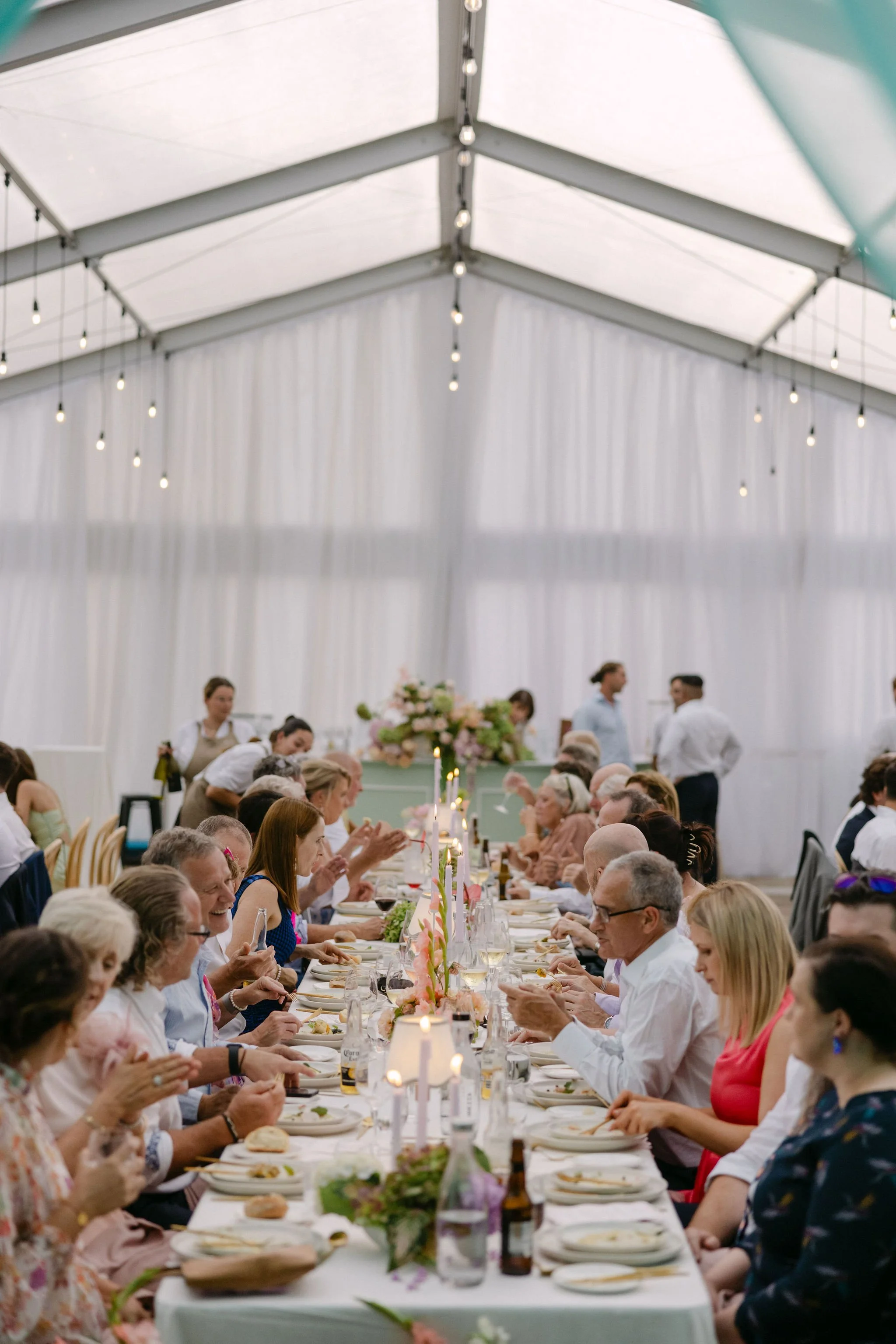 Guests dining at a long table decorated with flowers and candles beneath a white marquee and string lights.
