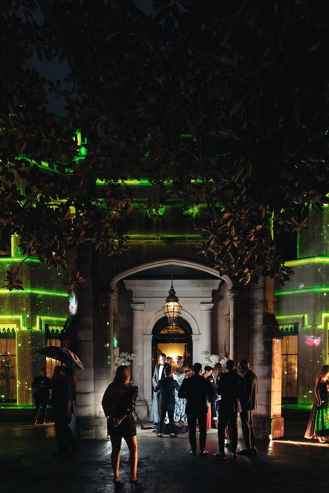 People gathered outside a building at night, with some standing under an umbrella, illuminated by lights and surrounded by dark trees and greenery.