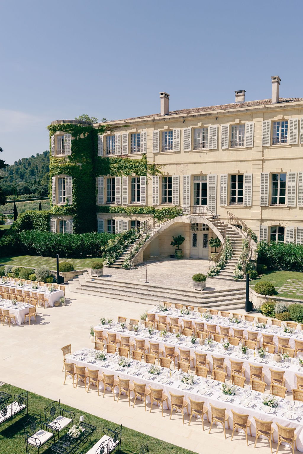 Outdoor wedding setup with long dining tables adorned with white tablecloths and floral centerpieces, in front of a large, elegant mansion with white shutters and lush greenery.