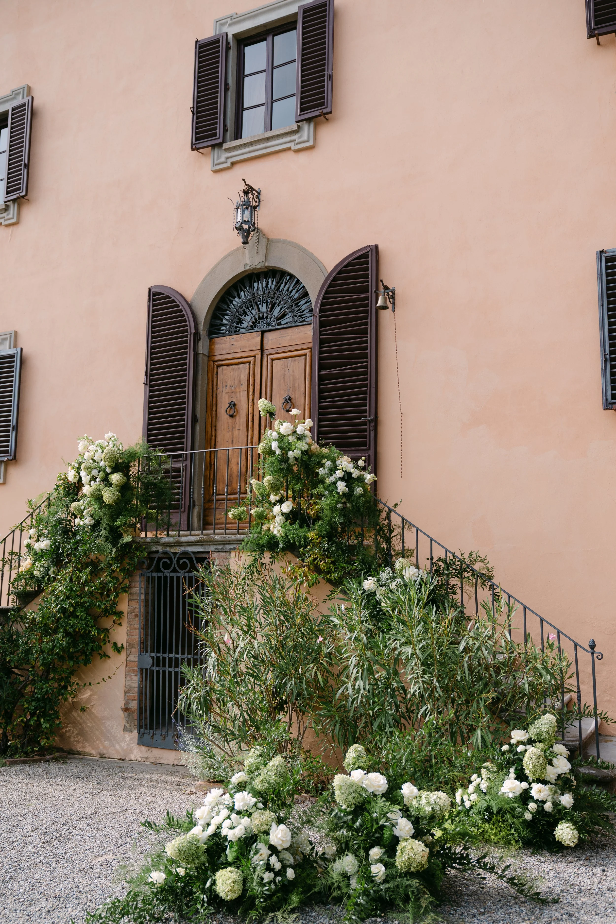 A peach-colored house with dark brown shutters, a wooden front door, and a small balcony. The house is decorated with lush white flowers and greenery, including hydrangeas and roses, near the entrance and along the stairs leading up to the door.