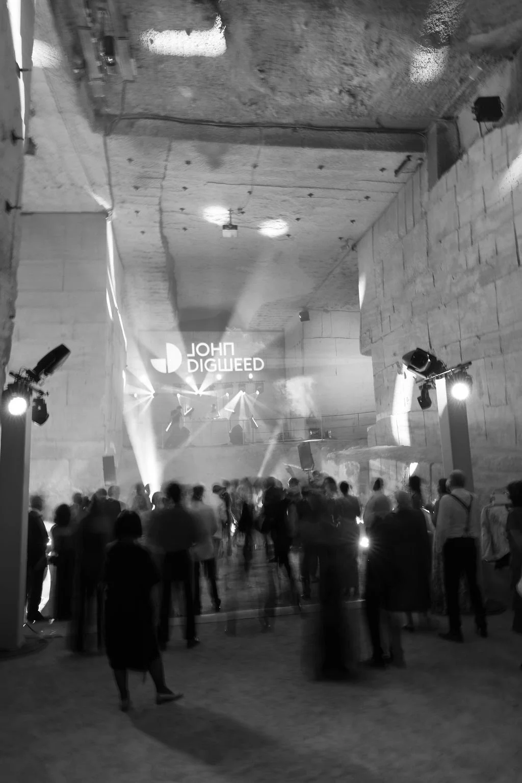 A large crowd of people gathers at an indoor concert or event with stage lighting and fog effects. A large screen displays the name 'John Digweed'.