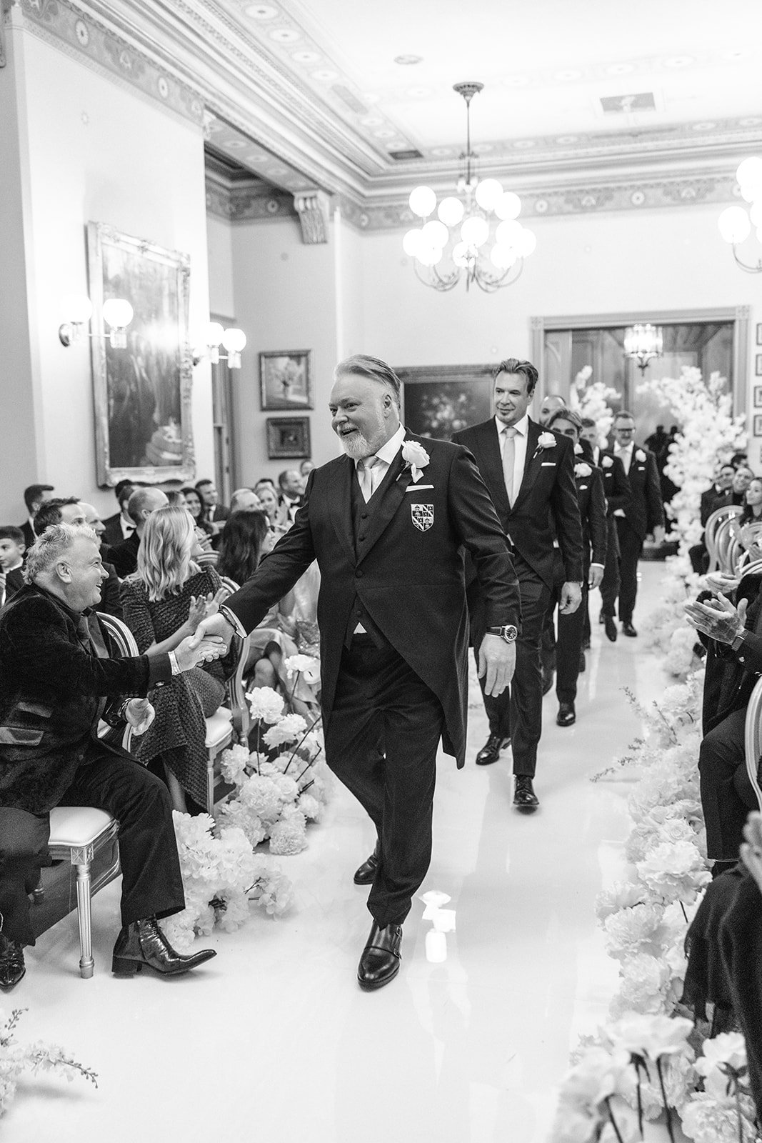 A black and white photo of a wedding ceremony where a group of men in tuxedos walks down the aisle, shaking hands with guests seated on either side. The setting appears to be an elegant, decorated hall.