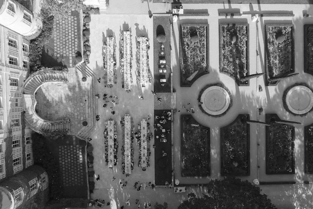 An aerial view of an outdoor area with tables, chairs, a water fountain, and garden beds, surrounded by buildings.