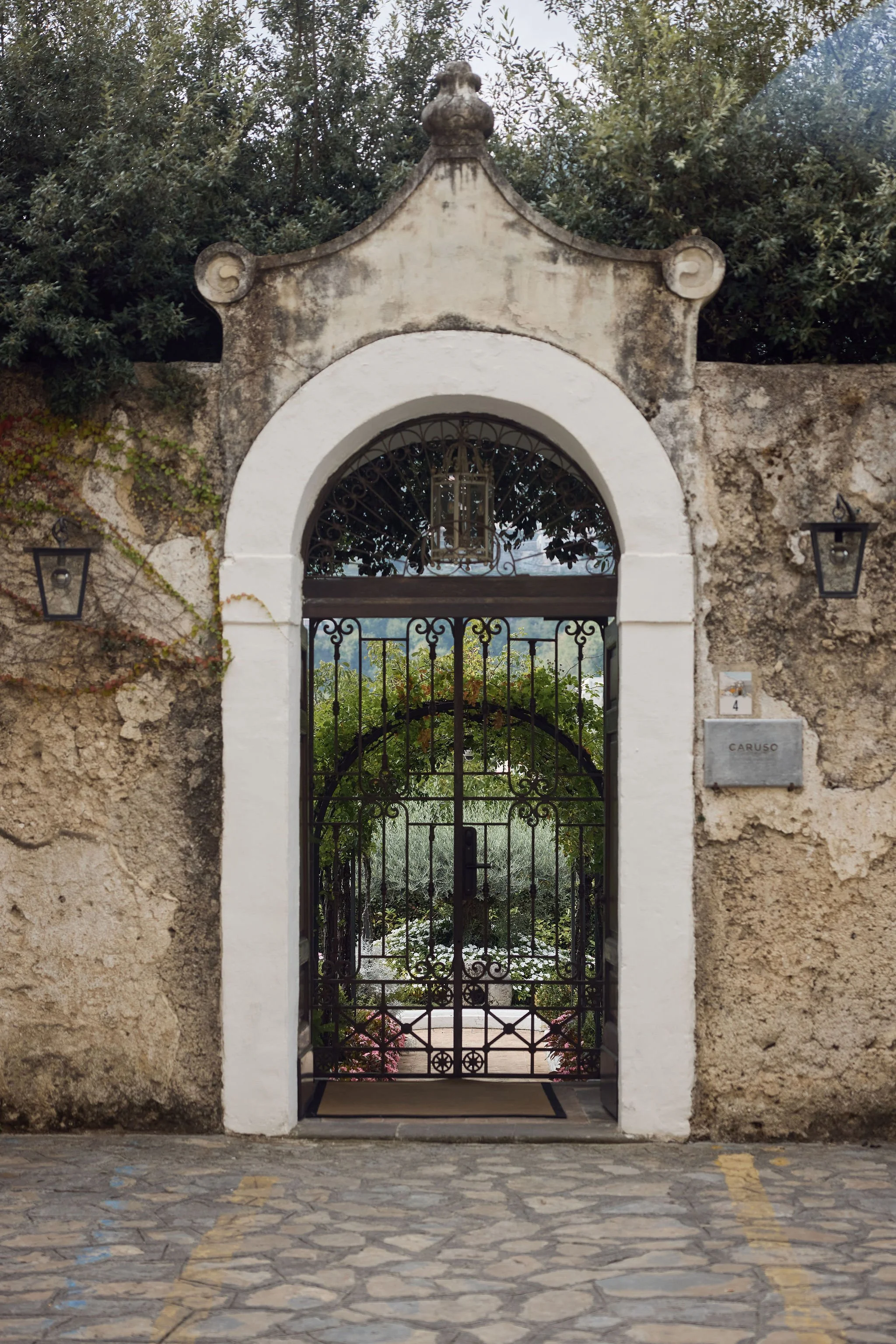 A wrought iron gate set in an arched stone entryway leading to a garden with greenery and flowers, with a rustic wall and lanterns on either side.