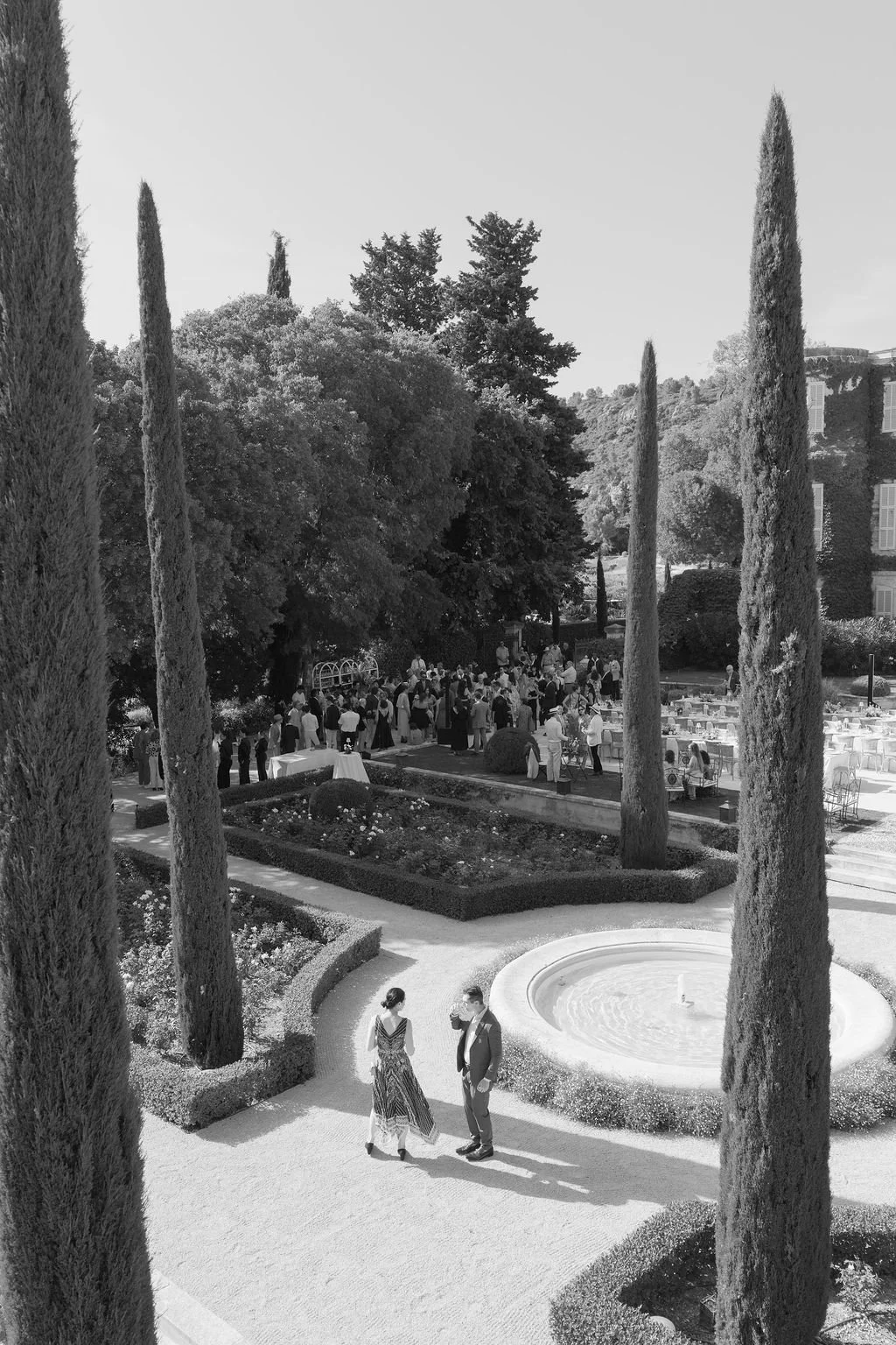 People attending an outdoor event in a formal garden with tall trees, flowers, a fountain, and white tables.