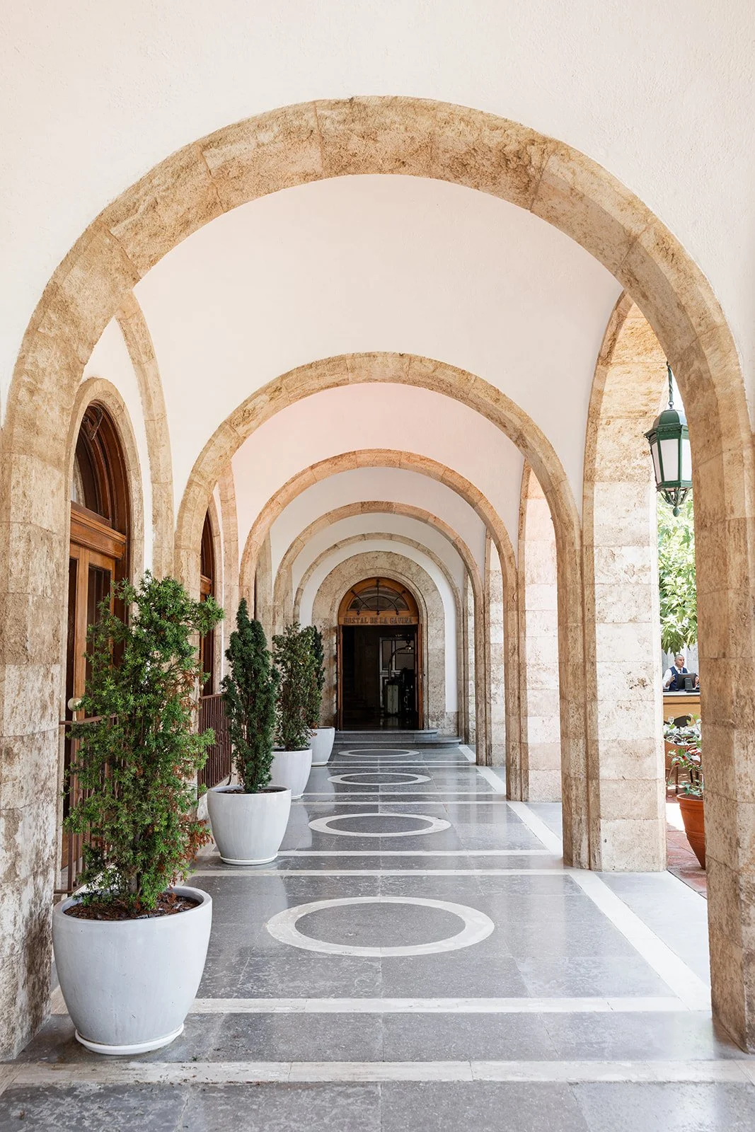A covered stone walkway with rounded arches, potted plants, and patterned flooring leads to a door.