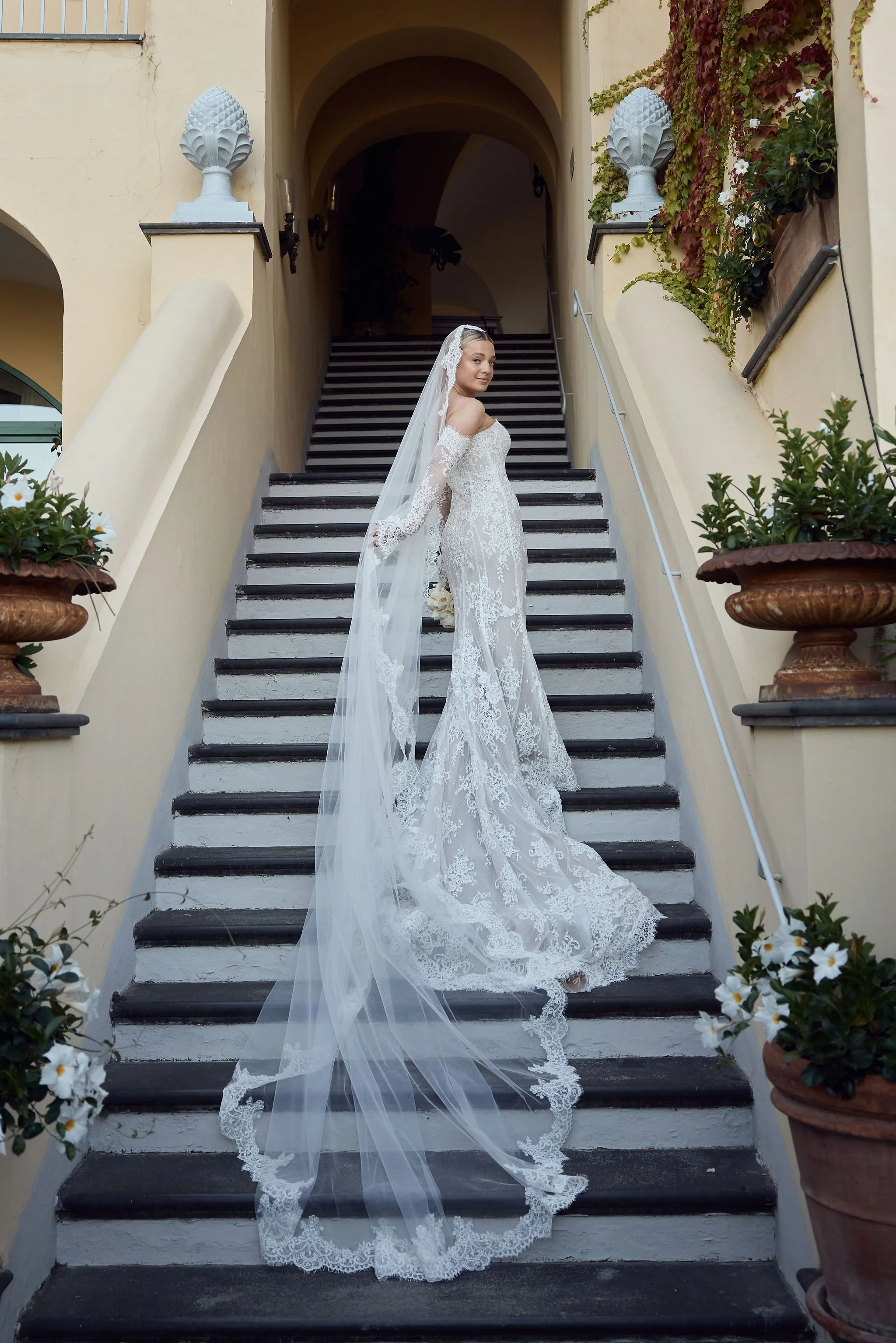 Bride in a white lace wedding gown with long train and veil standing on a staircase outside a building with potted plants.
