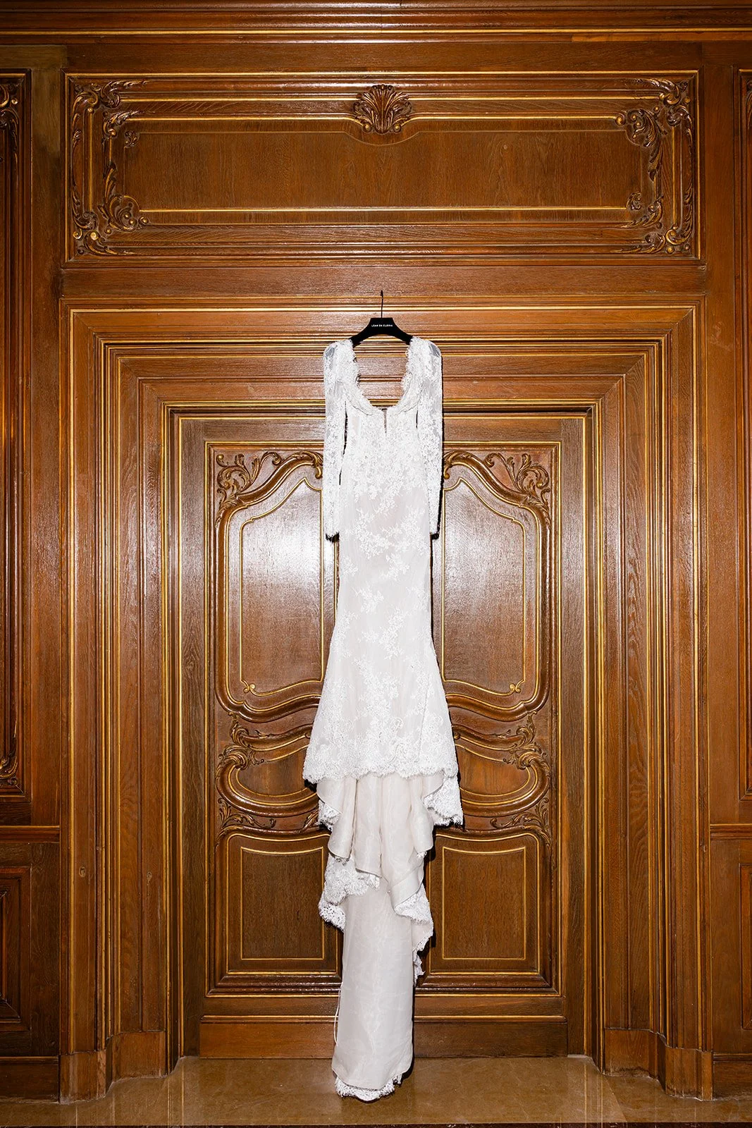 A white lace wedding dress hanging on a black hanger against a large, ornate wooden door with intricate carvings.