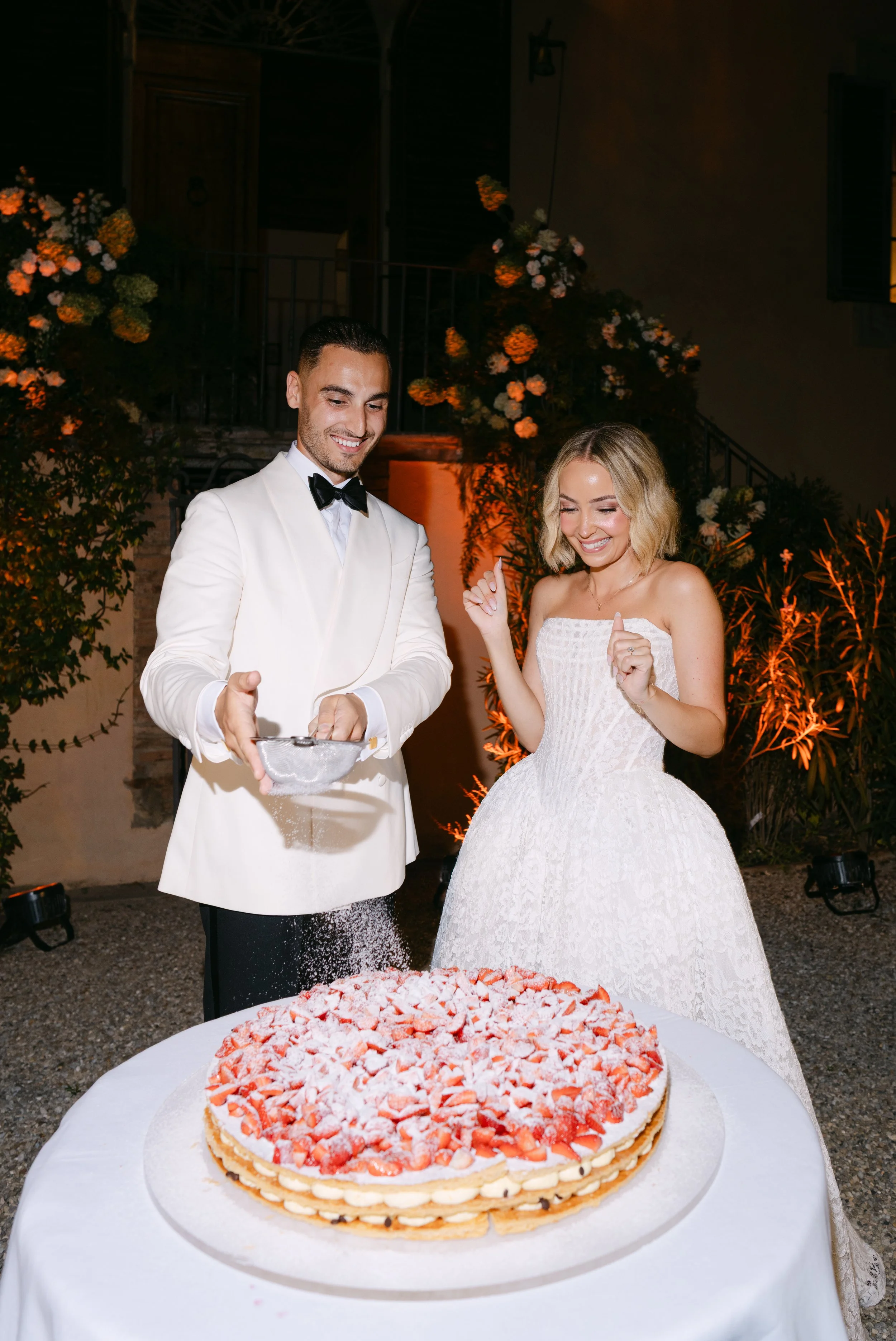 A bride and groom celebrating at their wedding reception with a strawberry cake on a table. The groom is wearing a white tuxedo with a black bow tie, and the bride is wearing a white wedding dress. They appear happy and are smiling, with the groom ho