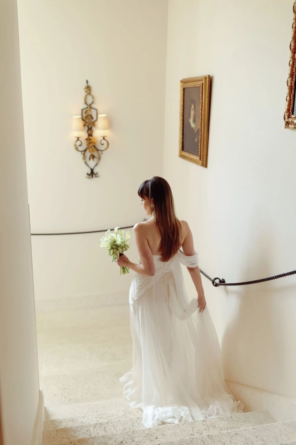 A woman in a white wedding dress holding a bouquet of white flowers, descending a staircase with cream-colored walls and ornate framed paintings.
