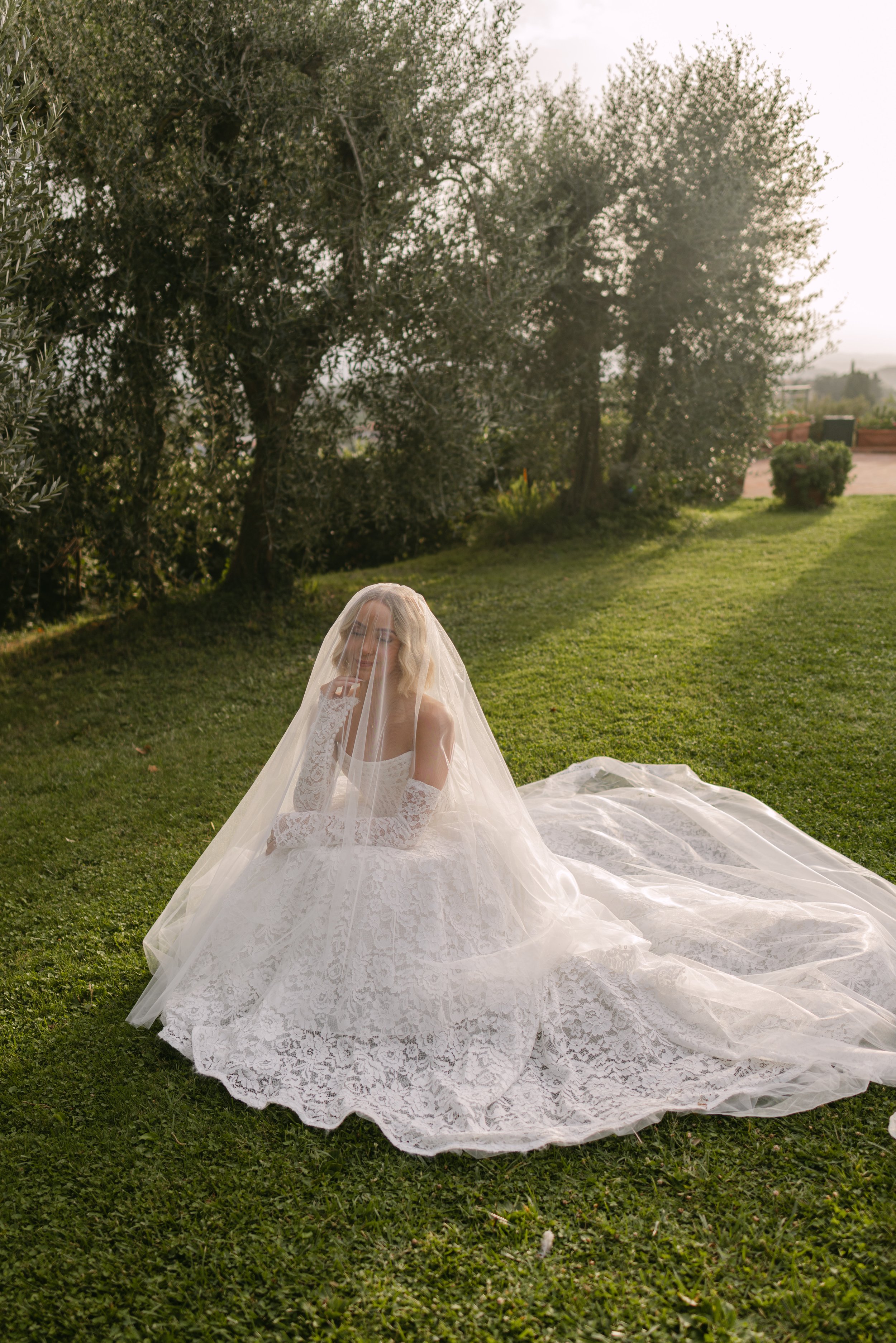 A woman dressed in a white lace wedding gown with a veil sitting on grass outside, surrounded by trees and greenery, during daylight.