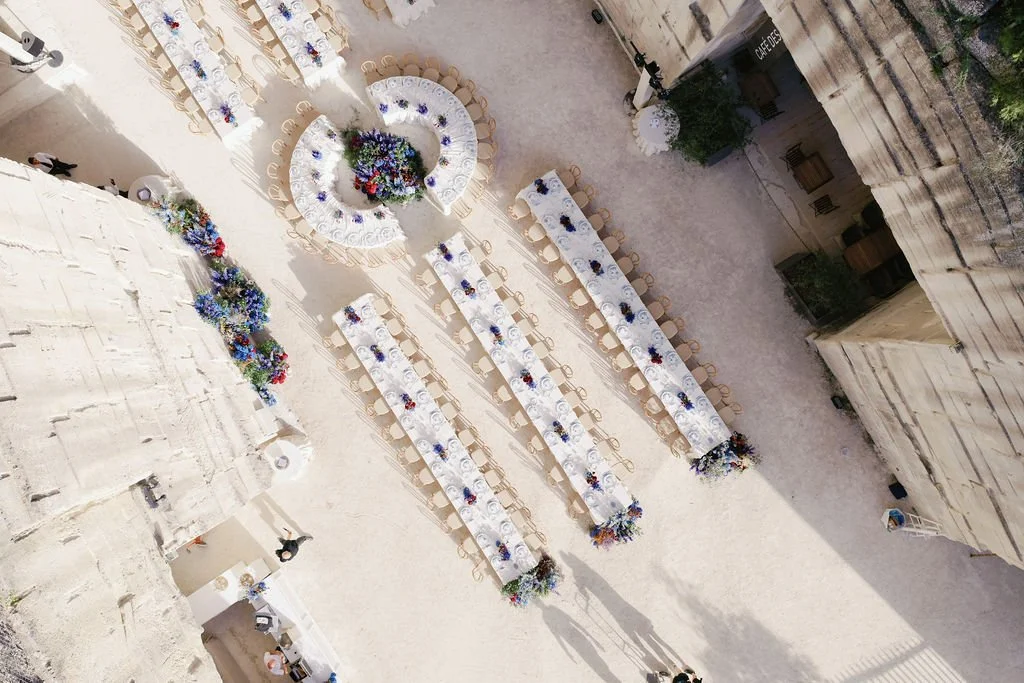 Outdoor event setup with long dining tables decorated with flowers, arranged in a courtyard with stone walls.
