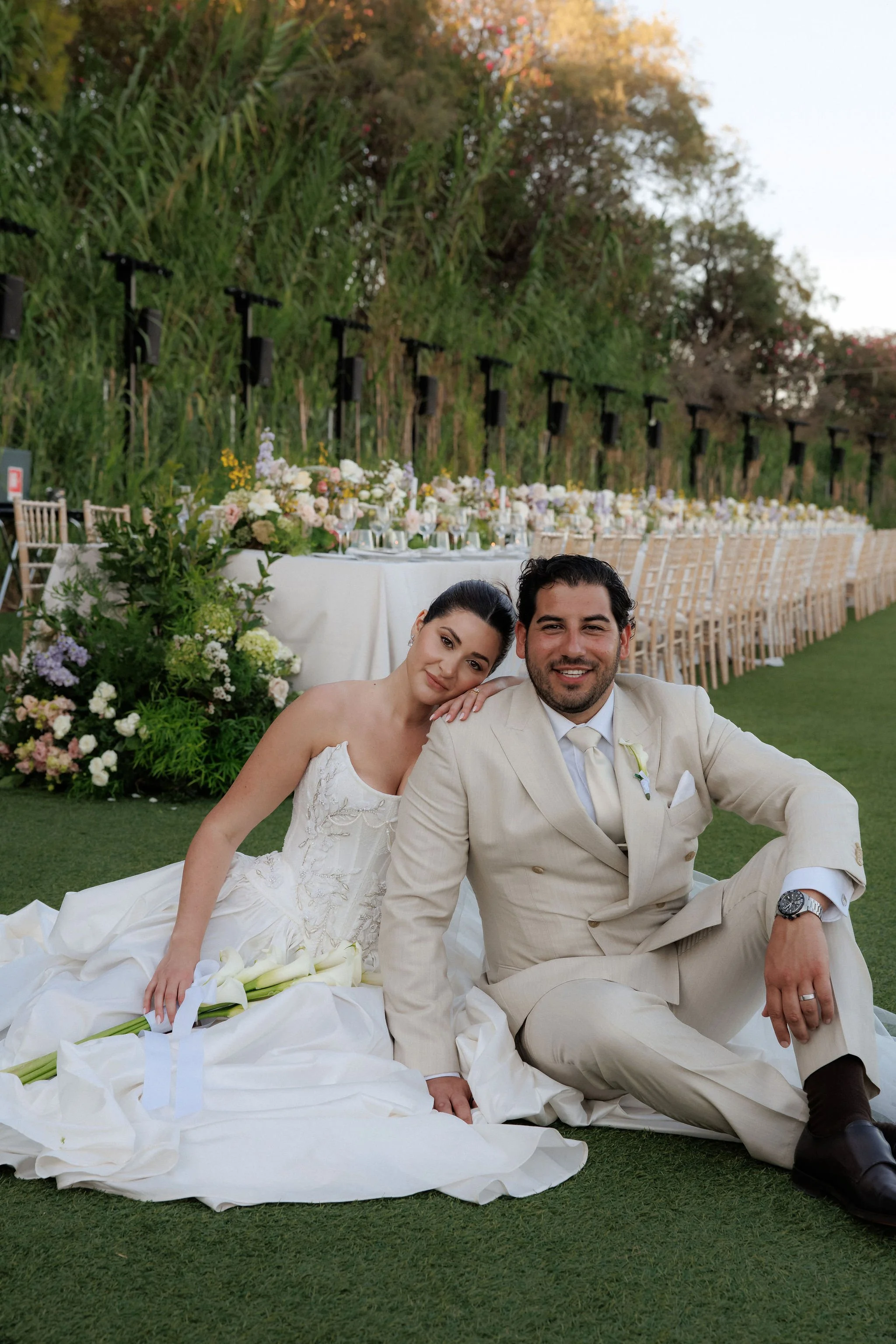 A bride and groom sitting on grass at their outdoor wedding reception, surrounded by floral arrangements and long banquet tables.