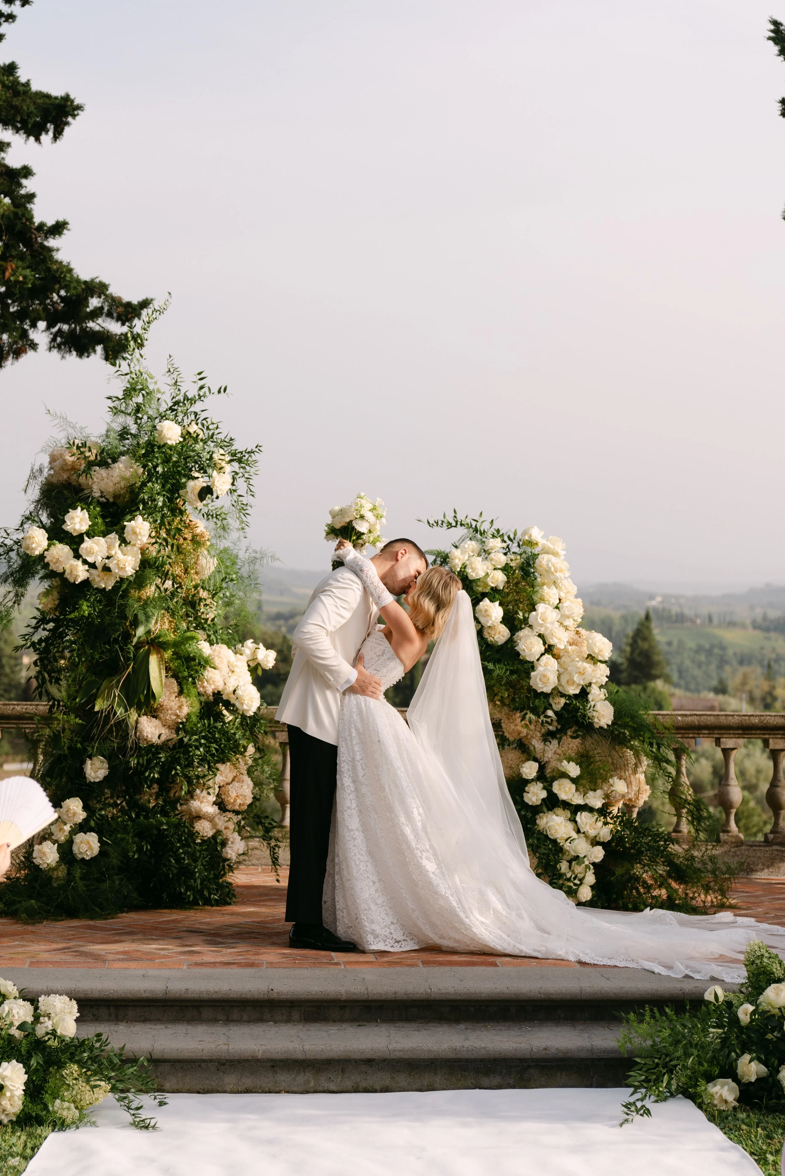 A bride and groom kiss during their wedding ceremony outdoors on a terrace with a scenic view, decorated with white and cream flowers and greenery.