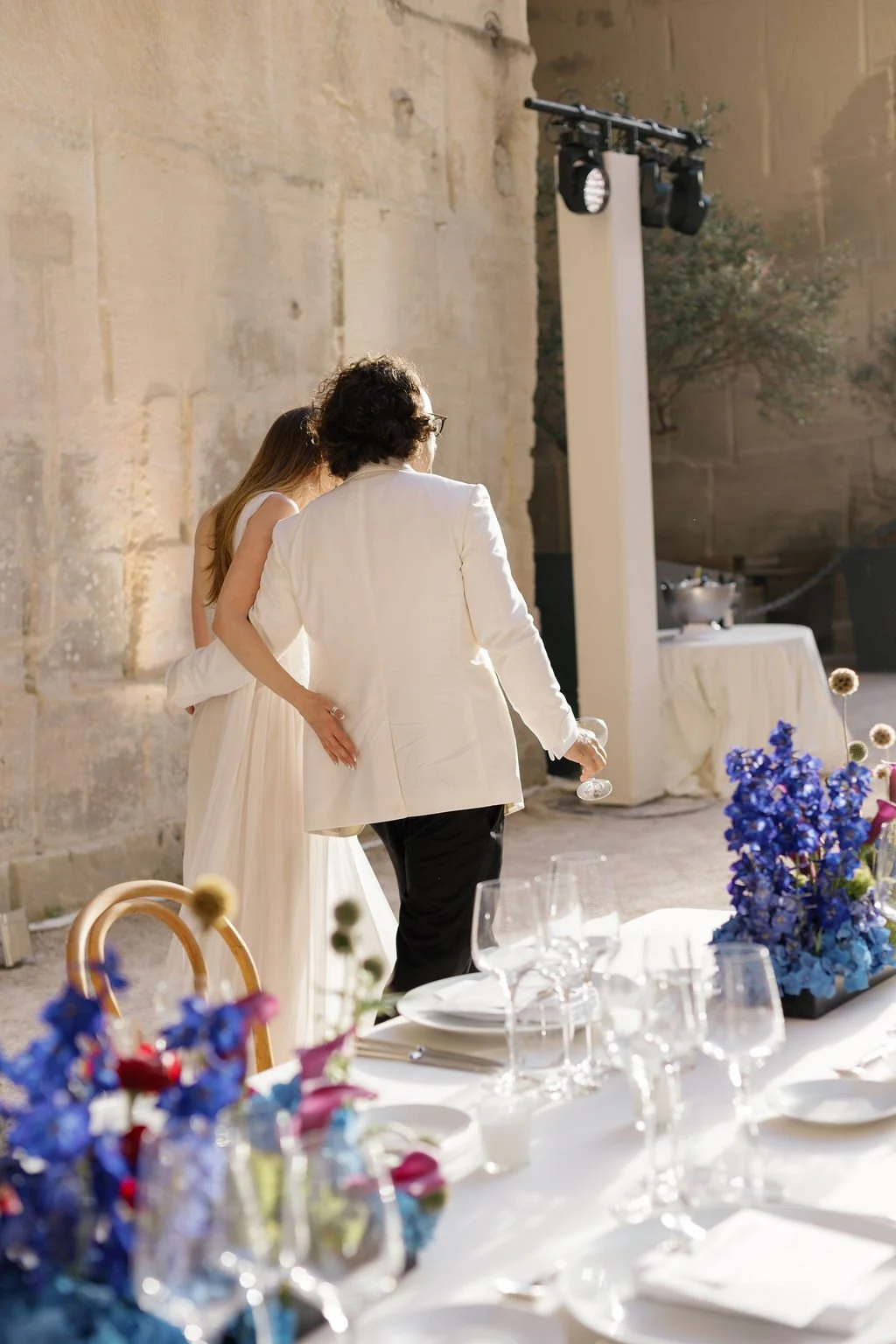 People at a wedding reception, with a woman in a white dress and a man in a white blazer and black pants, standing near a table decorated with flowers, holding glasses, in an outdoor setting with stone walls and sunlight.
