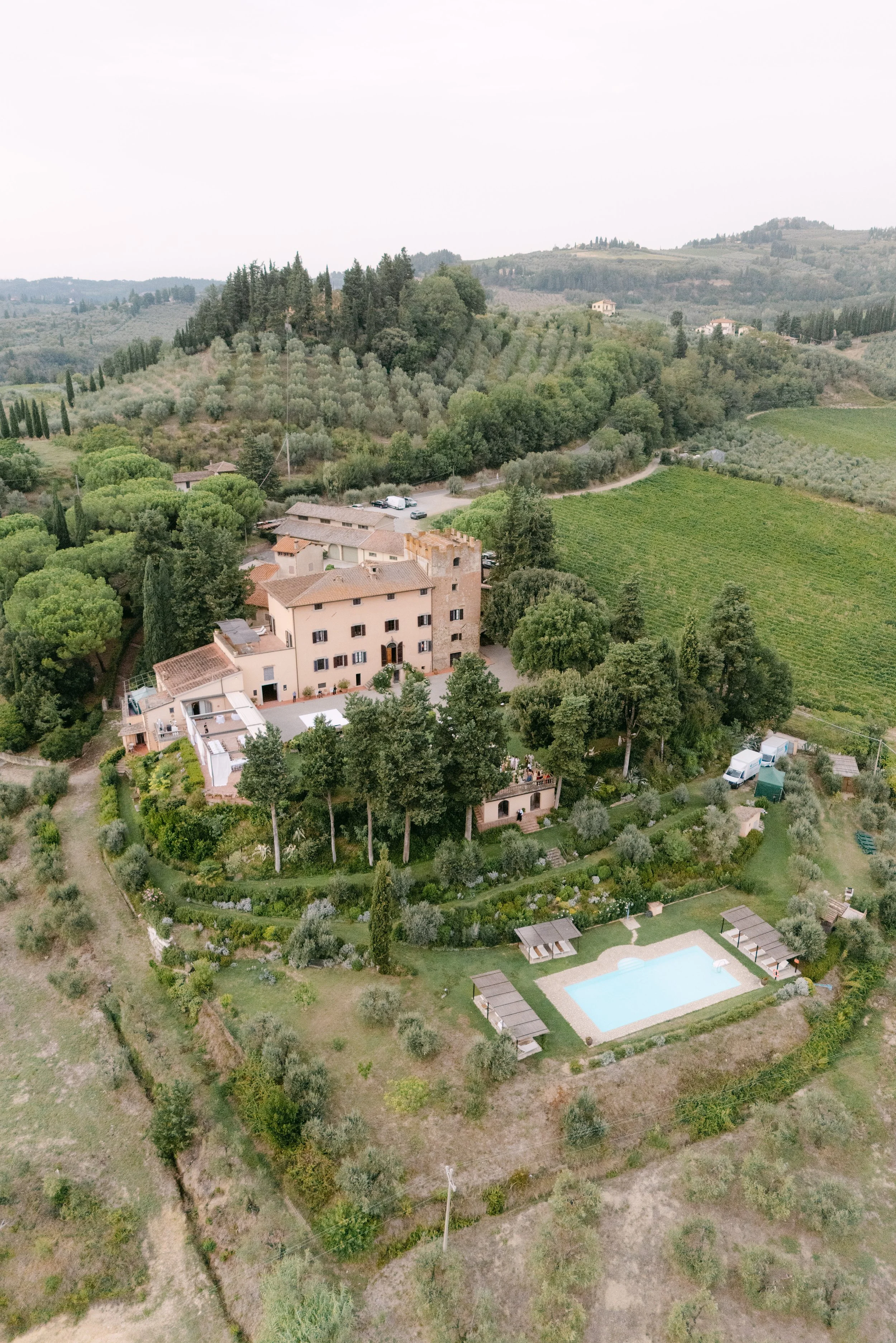A scenic landscape of a rural area with a large house, lush green trees, rolling hills, vineyards, and a swimming pool with lounge chairs.