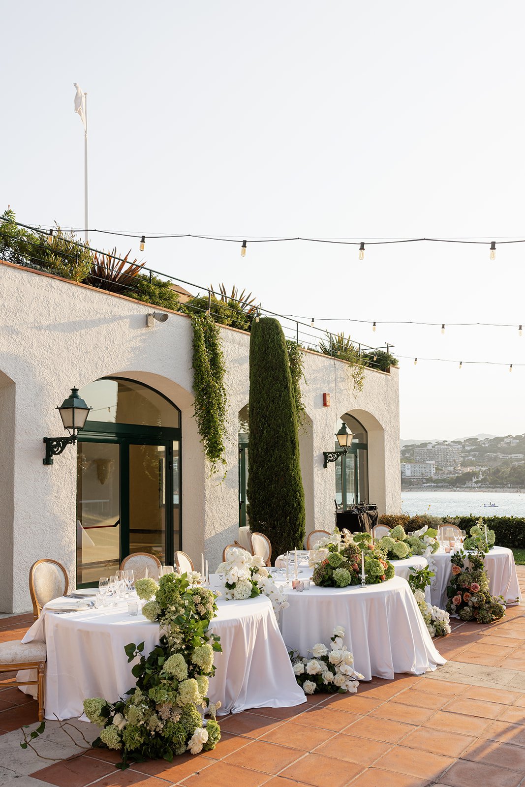 Outdoor event setup with long tables decorated with white tablecloths and floral arrangements, overlooking water with a white building on one side, trees, and string lights overhead.