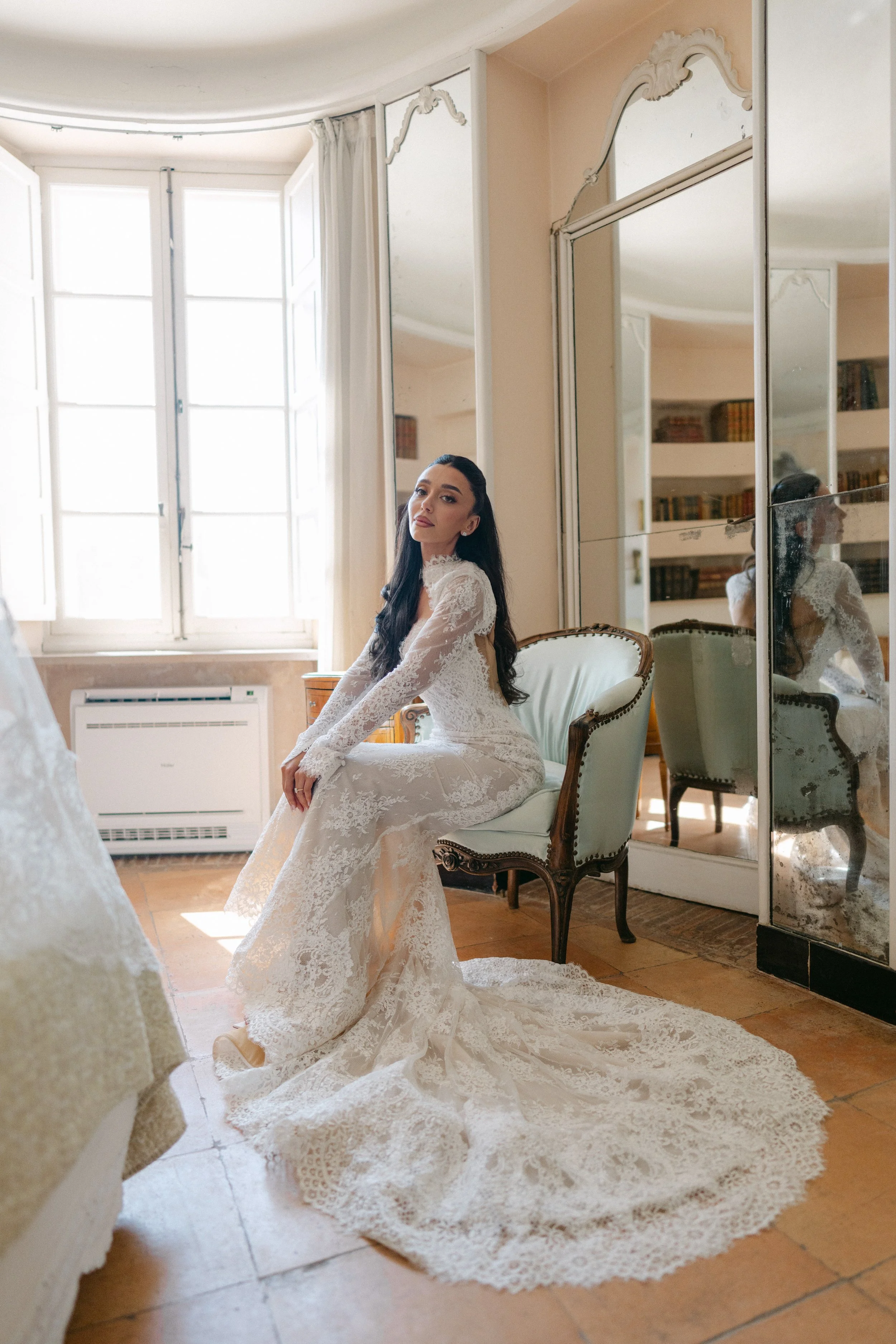 A woman in an elegant white lace wedding dress poses sitting on a vintage armchair in a well-lit room with large windows, mirrored closet doors, and a bookshelf in the background.