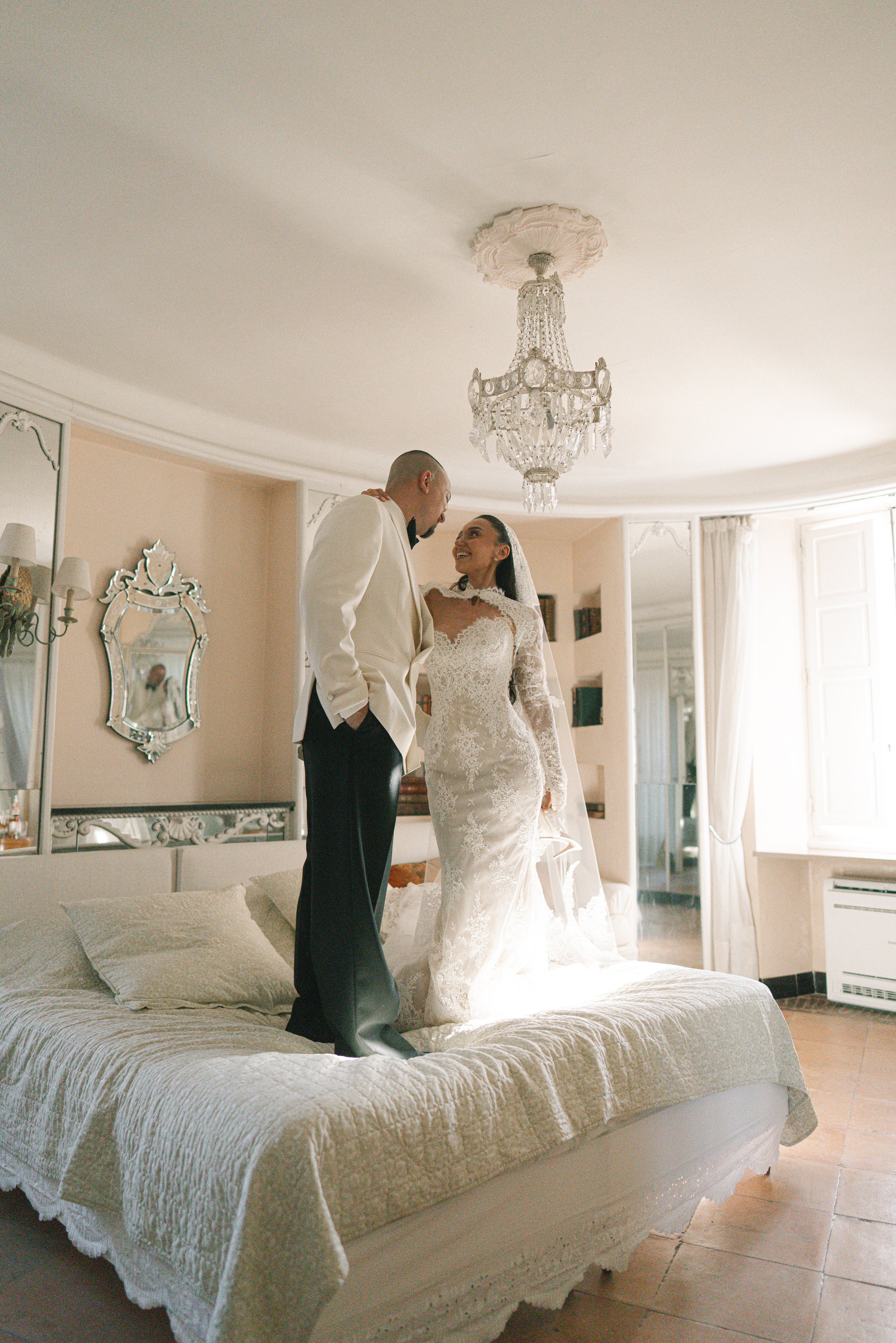 A bride and groom standing on a bed inside a well-lit room, smiling at each other. The bride is wearing a lace wedding gown and veil, and the groom is in a white tuxedo with black pants. The room has a chandelier hanging from the ceiling, and a mirro