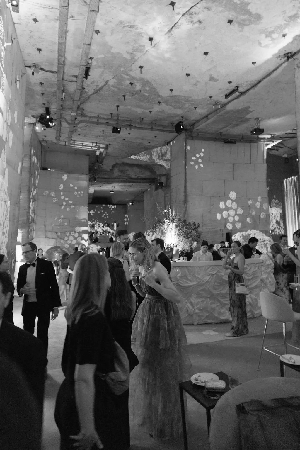 People dressed in formal attire socializing at a black-and-white indoor event with decorative lighting and floral arrangements.