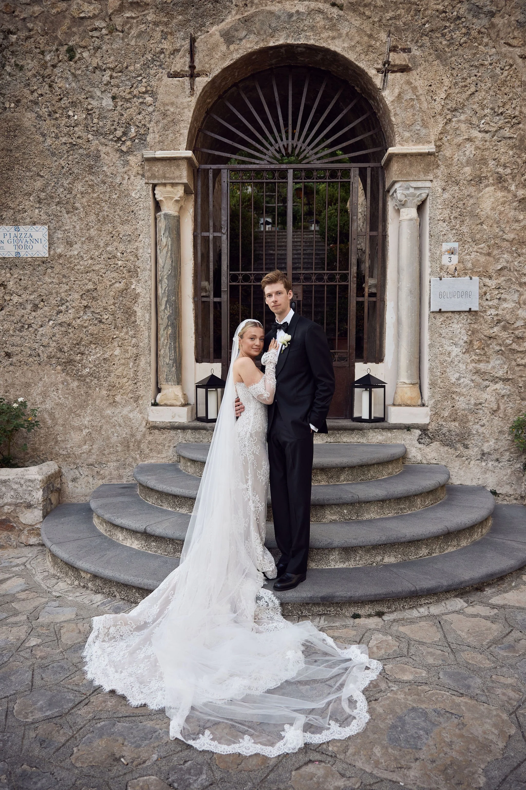 Bride and groom in wedding attire standing on stone steps in front of a gated archway outside a church or historic building.