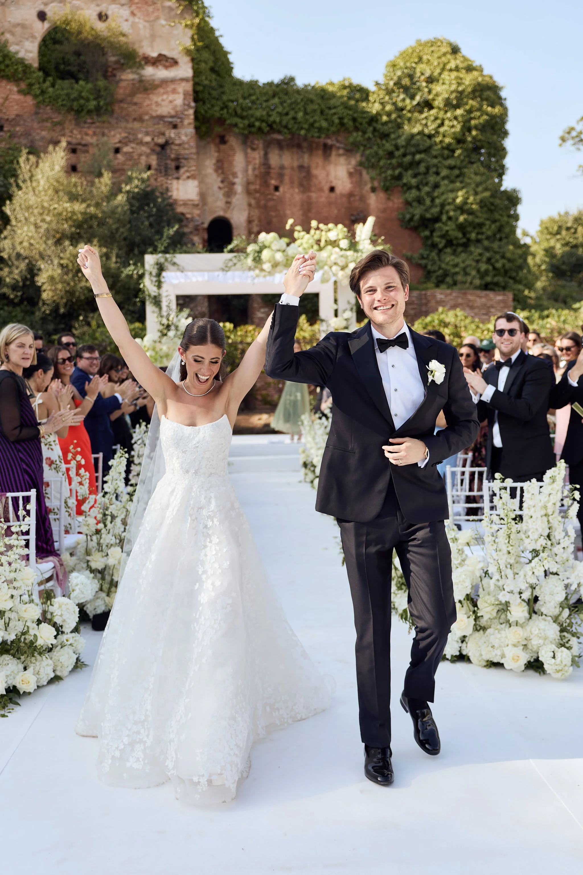 Newlyweds celebrate as they walk down a white flower-lined aisle.