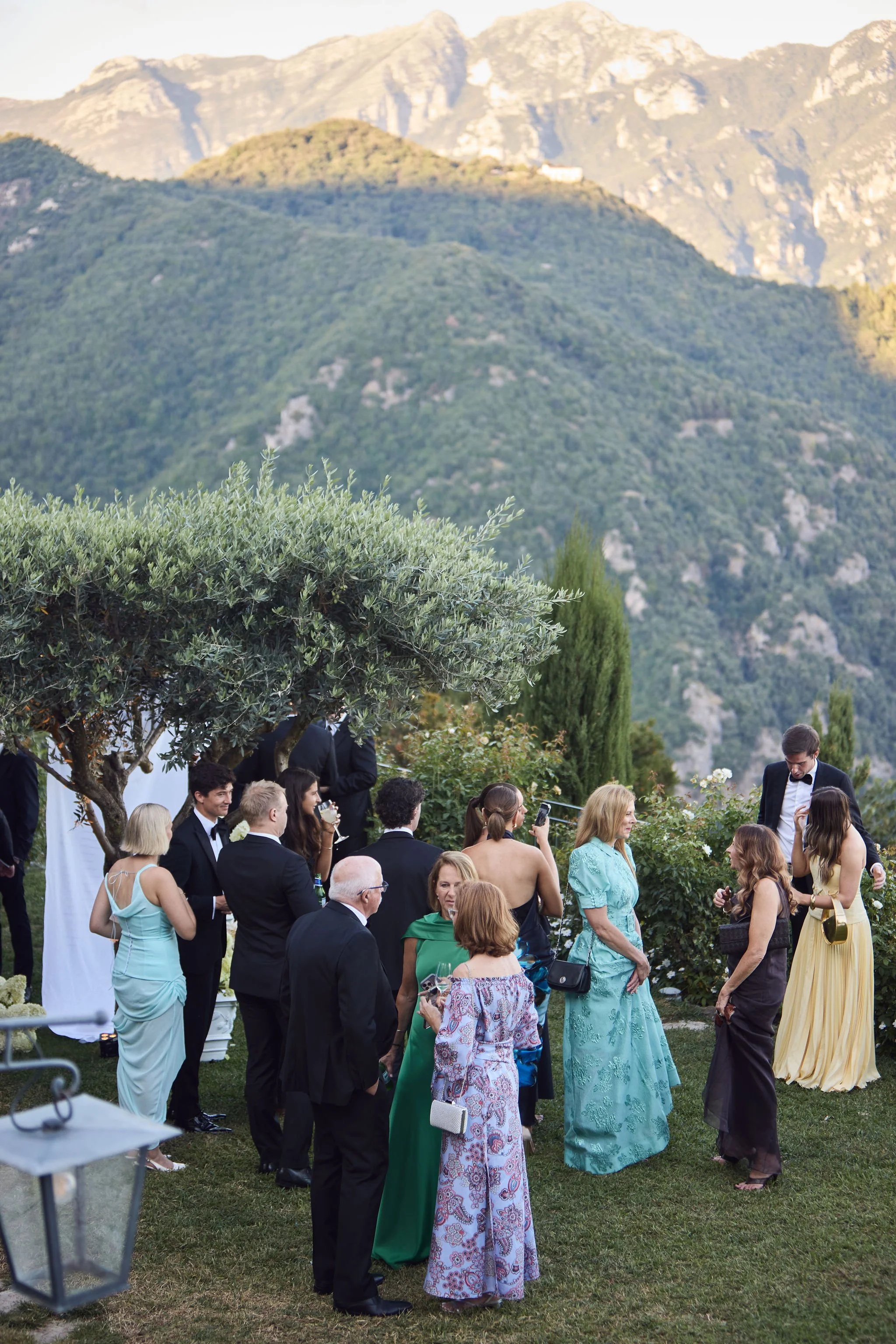 Group of elegantly dressed people socializing outdoors in a garden with mountains in the background.
