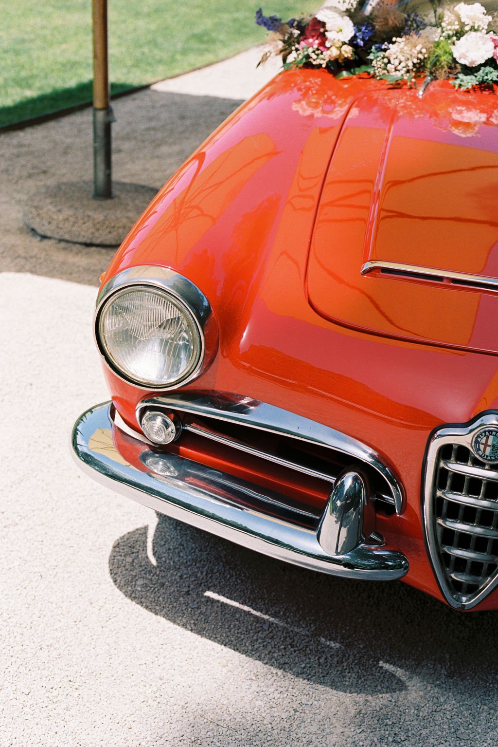 Close-up of the front of a vintage orange car decorated with a flower arrangement on the hood, featuring a prominent headlight, chrome details, and distinctive grille.