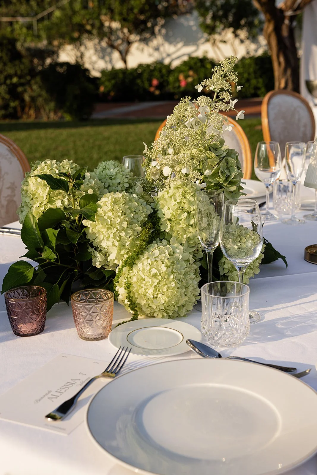 Elegant outdoor dining table with white tablecloth, floral centerpiece of white hydrangeas and greenery, surrounded by wine glasses, tea lights, and fine china, set in a garden setting