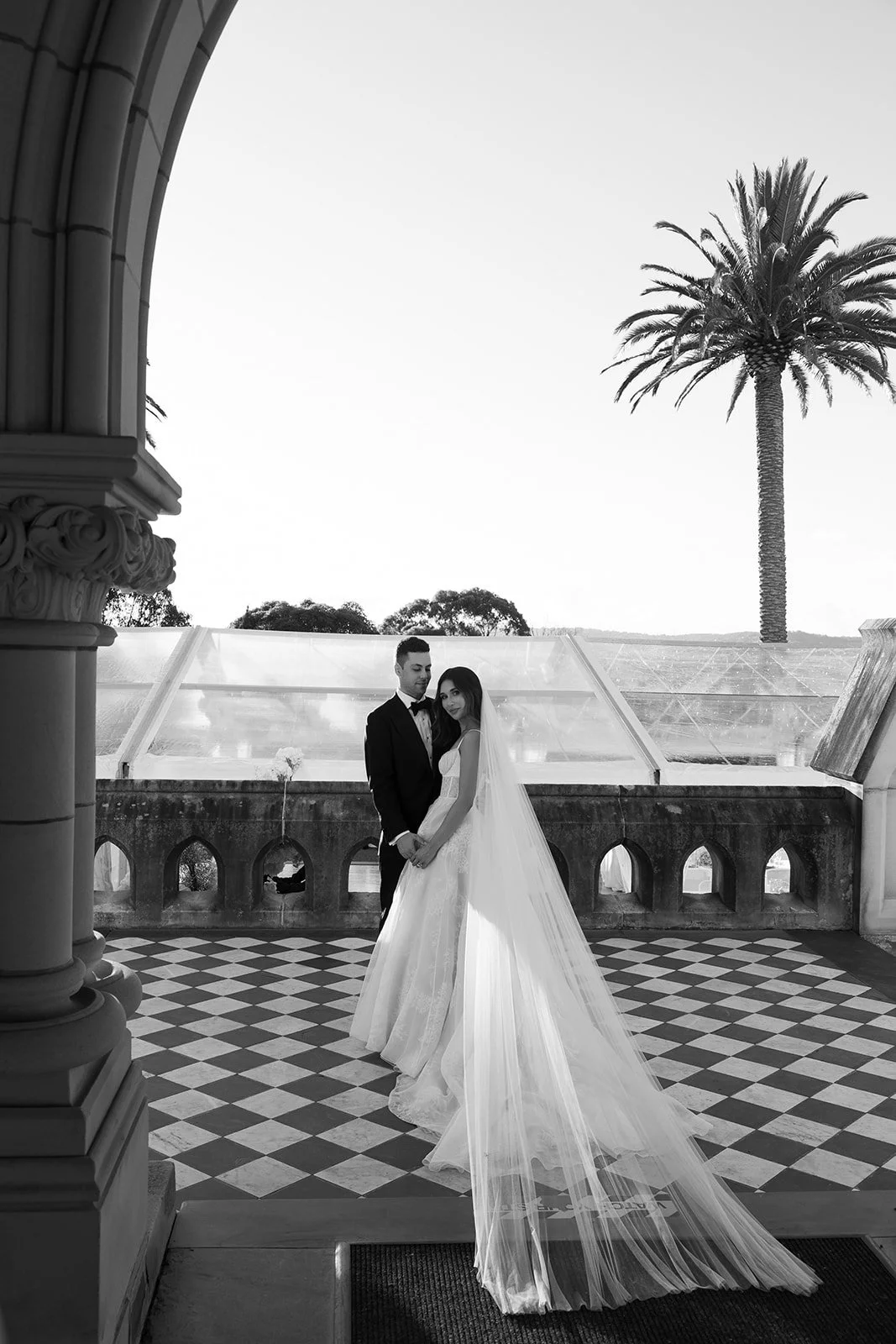Black and white photo of a bride and groom standing closely on a balcony with a checkered floor, palm tree, and a glass structure in the background.