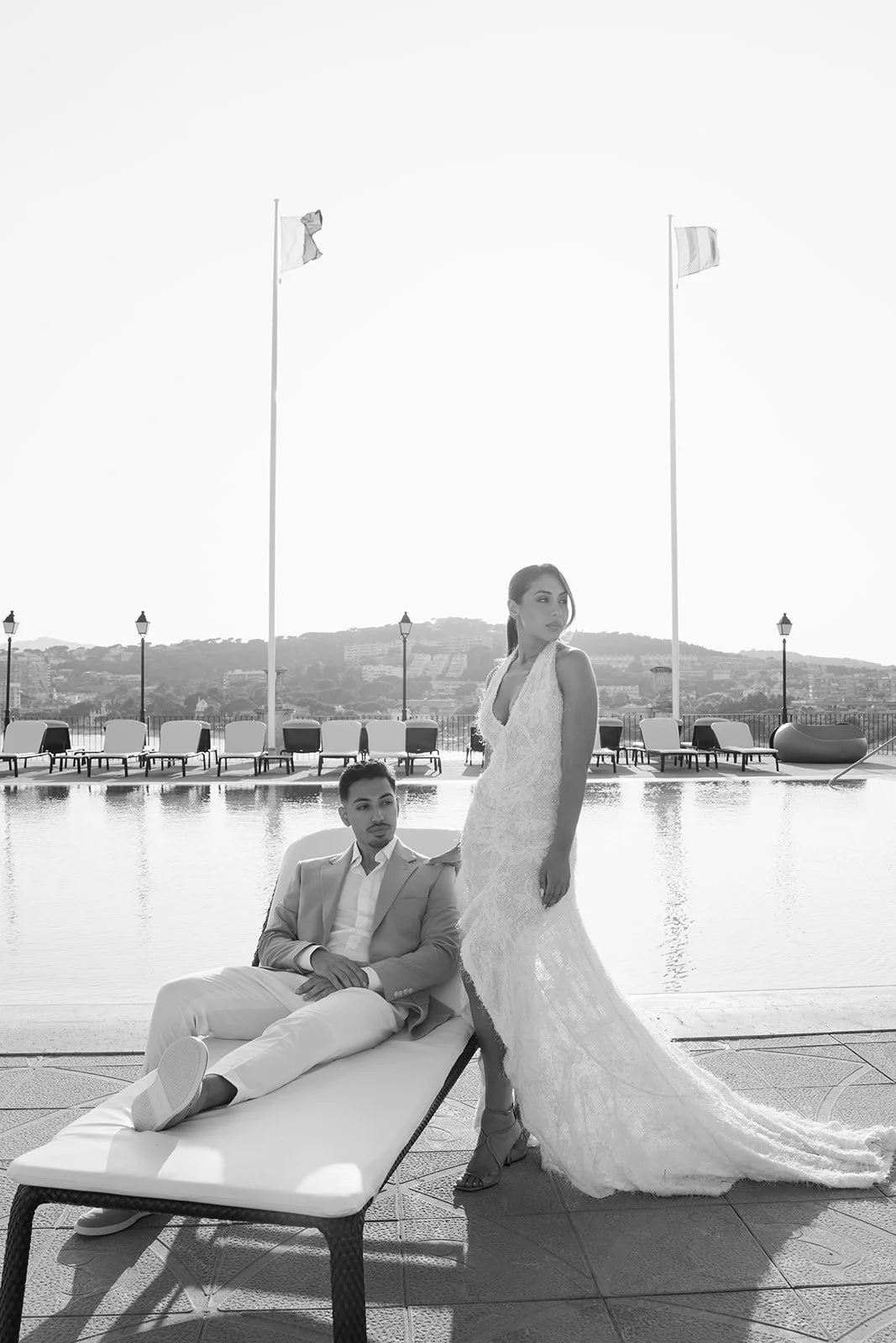 A black-and-white photo of a man in a light suit sitting on a lounger and a woman in a lace wedding dress standing beside him on a poolside deck with chairs, flags, and a distant cityscape in the background.