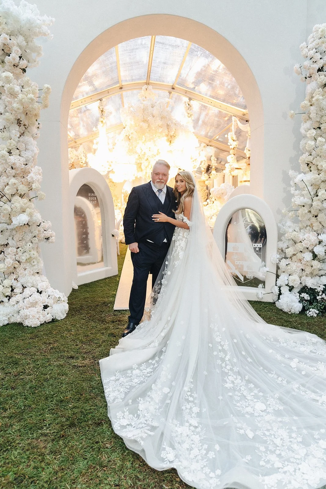 A bride in a white wedding gown with floral lace details standing beside a man in a dark suit, set in a decorated outdoor wedding venue with white floral arrangements and a lit canopy ceiling.
