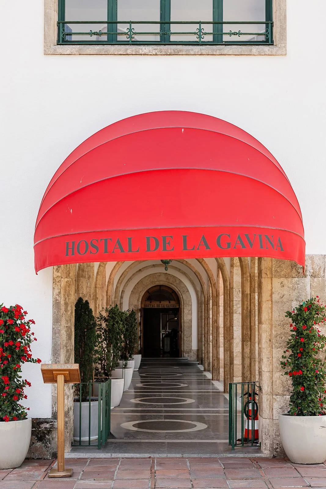 Entrance of a building with a red awning that reads "Hostal de la Gavina," with potted plants on each side and a patterned walkway leading to a door.