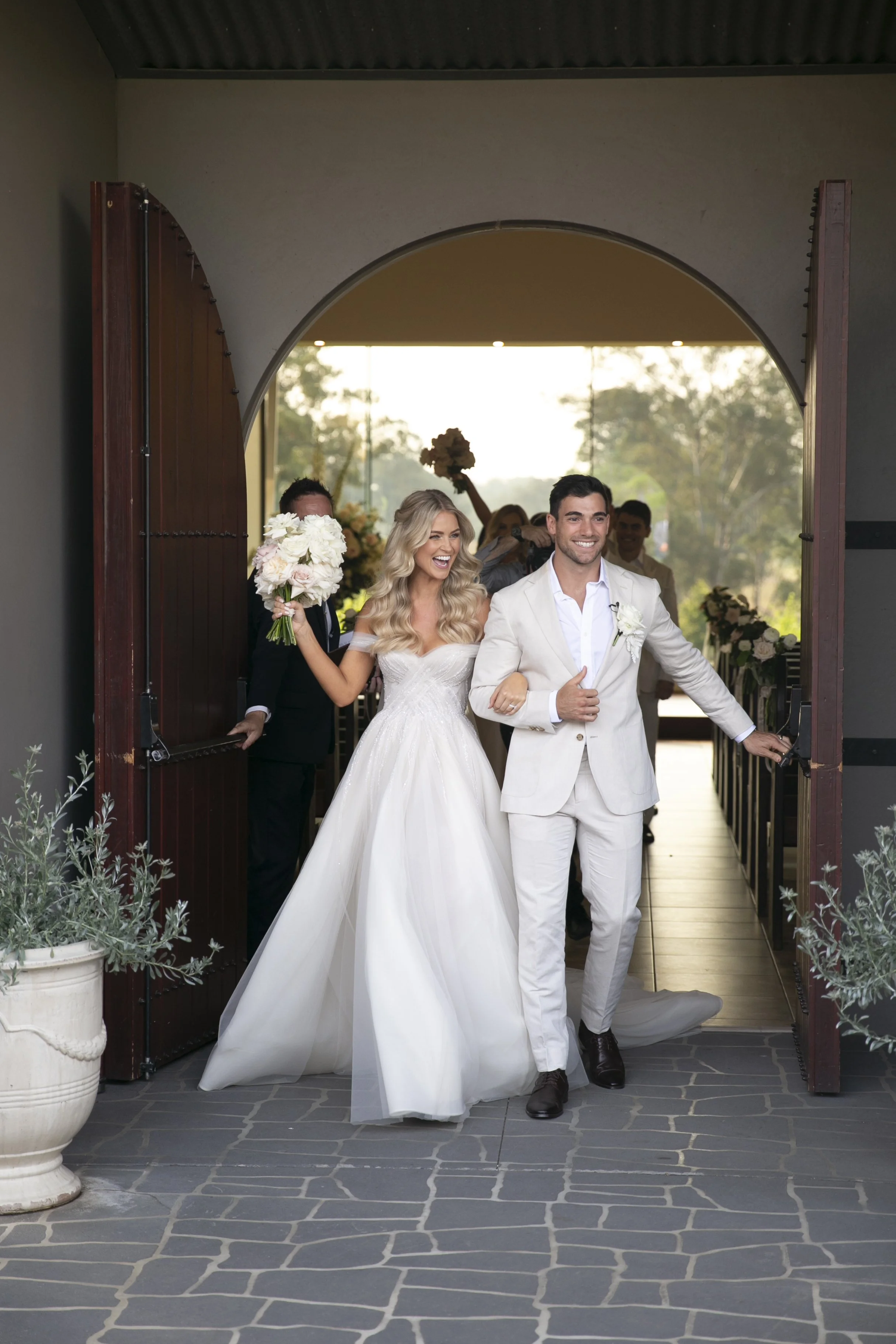A newlywed couple exits a building, smiling and celebrating, with the bride holding a bouquet of flowers and the groom dressed in a white suit. Guests are in the background holding flowers and celebrating.