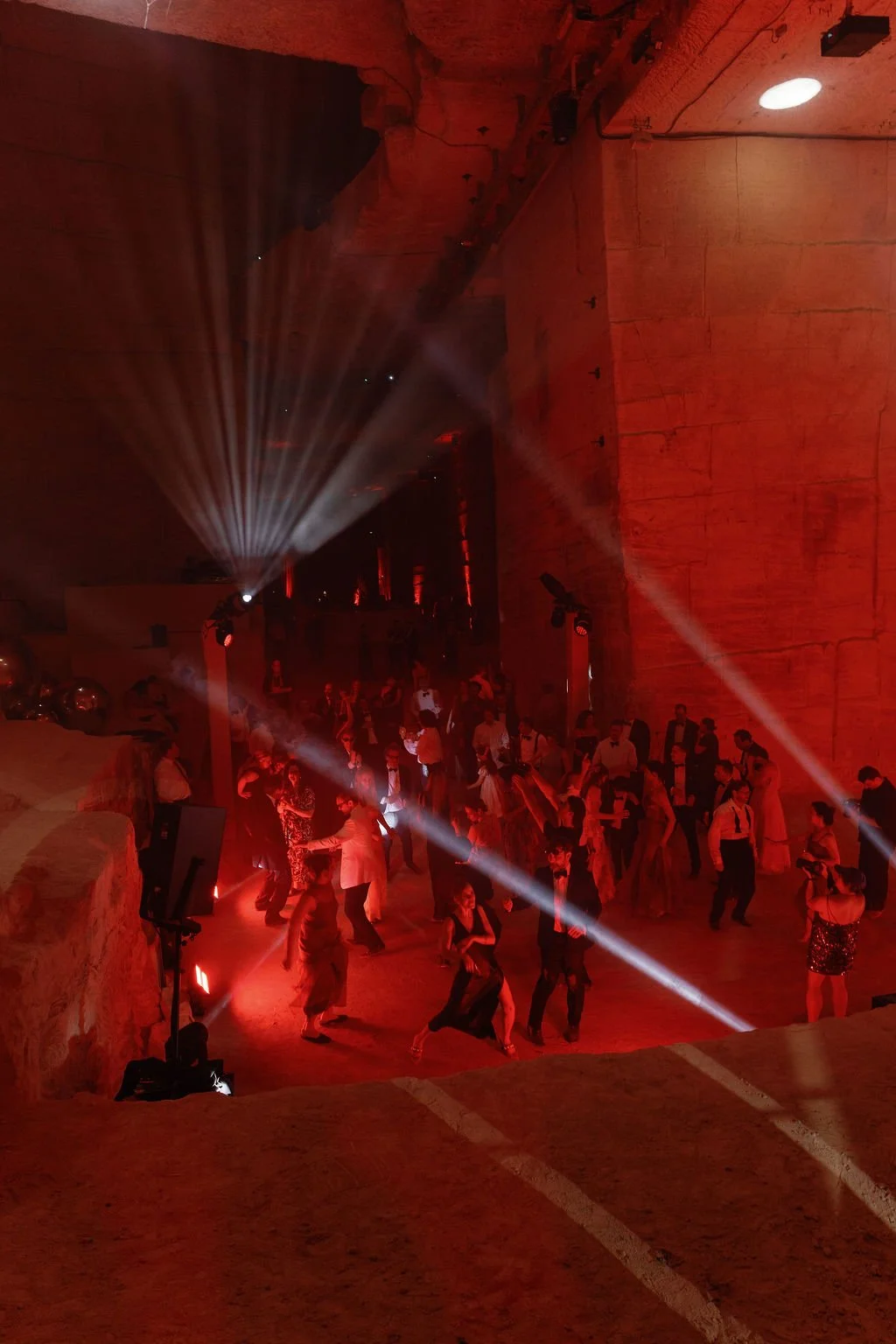 People dancing at a party or event beneath red lighting with spotlights and a large stone or concrete wall.