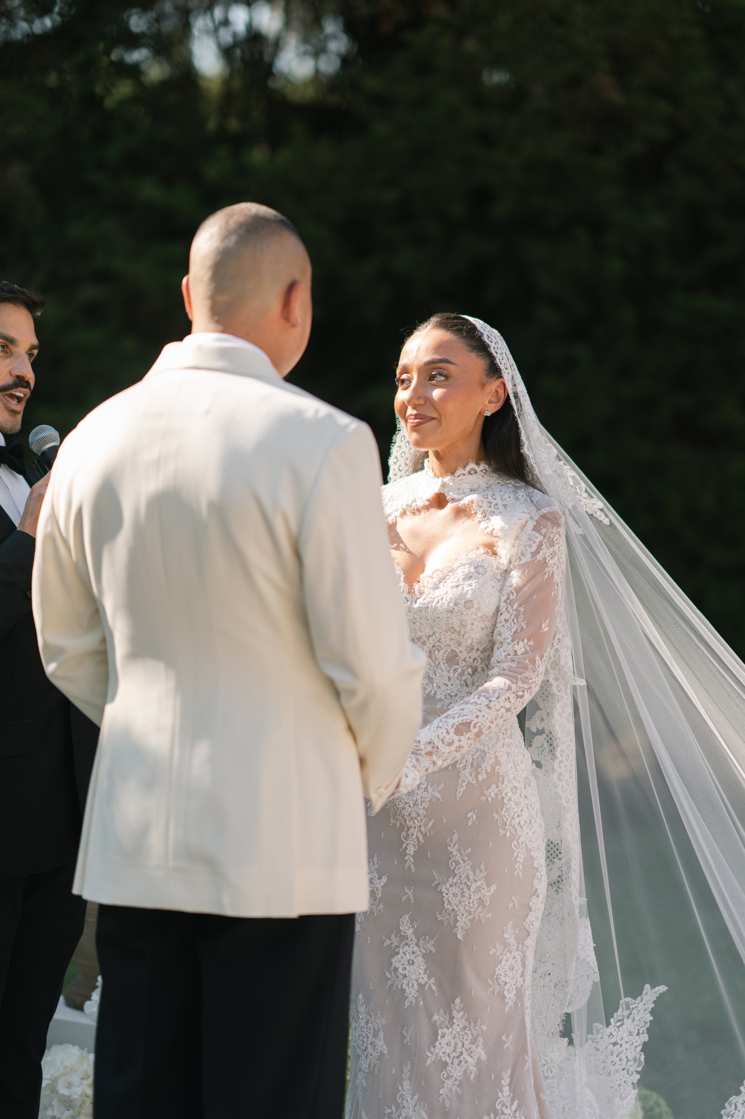 A bride and groom holding hands during outdoor wedding ceremony, bride wearing lace wedding dress with veil, groom in cream suit, officiant holding microphone on the left, green trees in background.