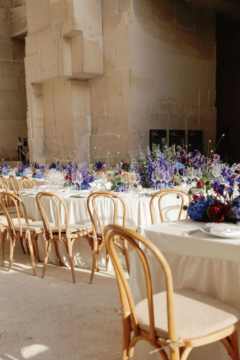 Elegant banquet hall with round tables draped in white tablecloths, decorated with purple and red floral centerpieces, wine glasses, and tableware, set against a backdrop of large stone wall at Picasso museum.