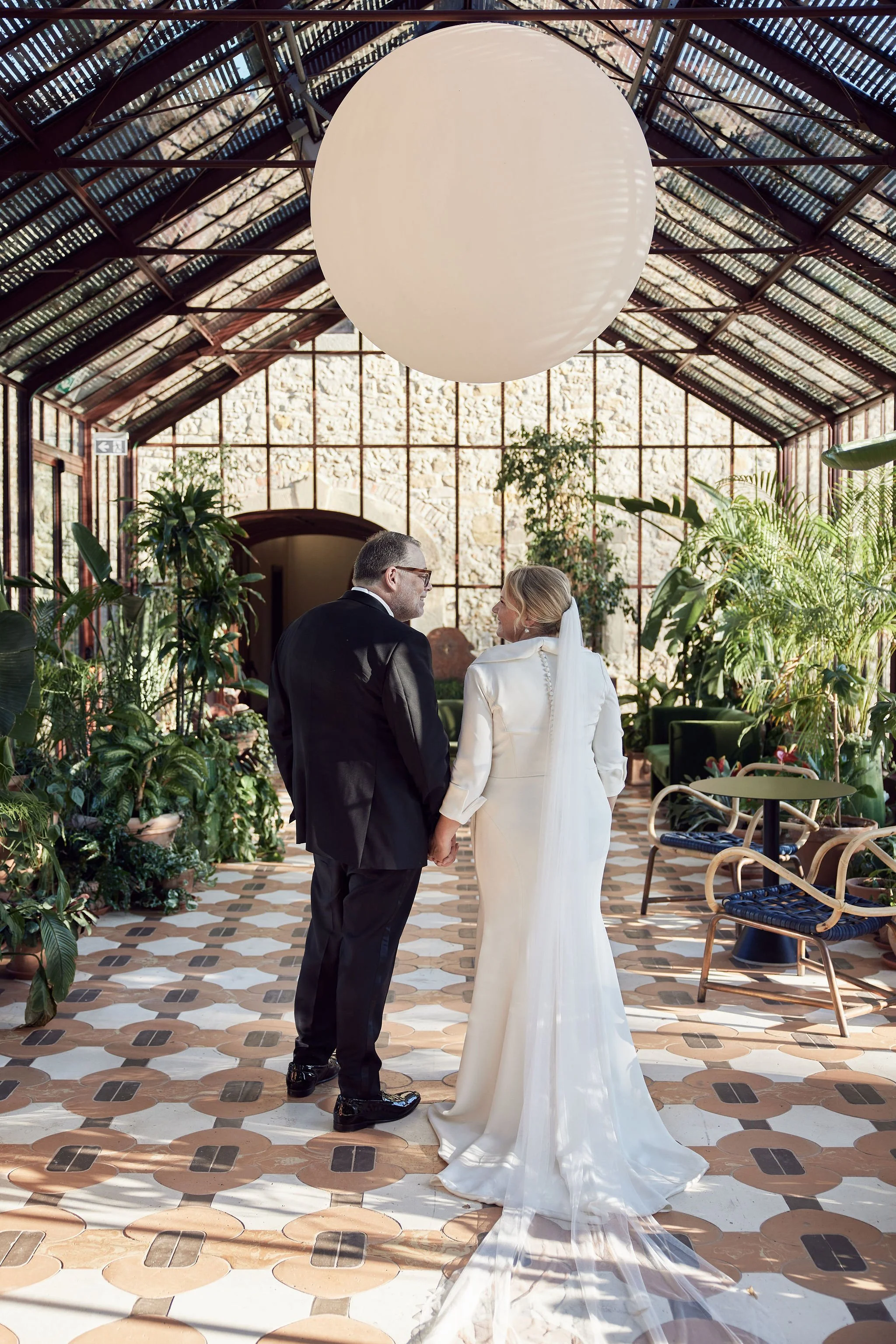 Bride and groom holding hands in a bright, plant-filled conservatory with a large spherical light fixture overhead.