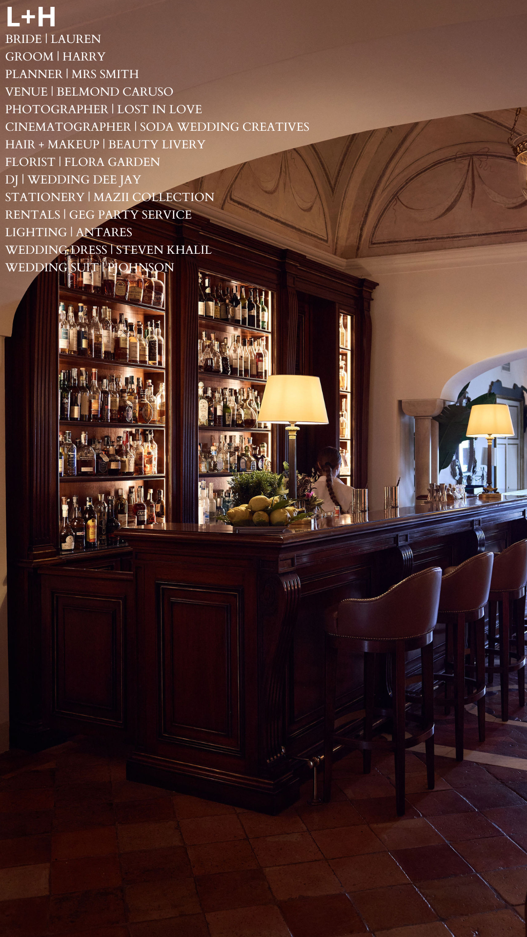 A cozy bar area with a wooden counter and barstools, illuminated by table lamps, with a large stock of liquor bottles on wooden shelves in the background.