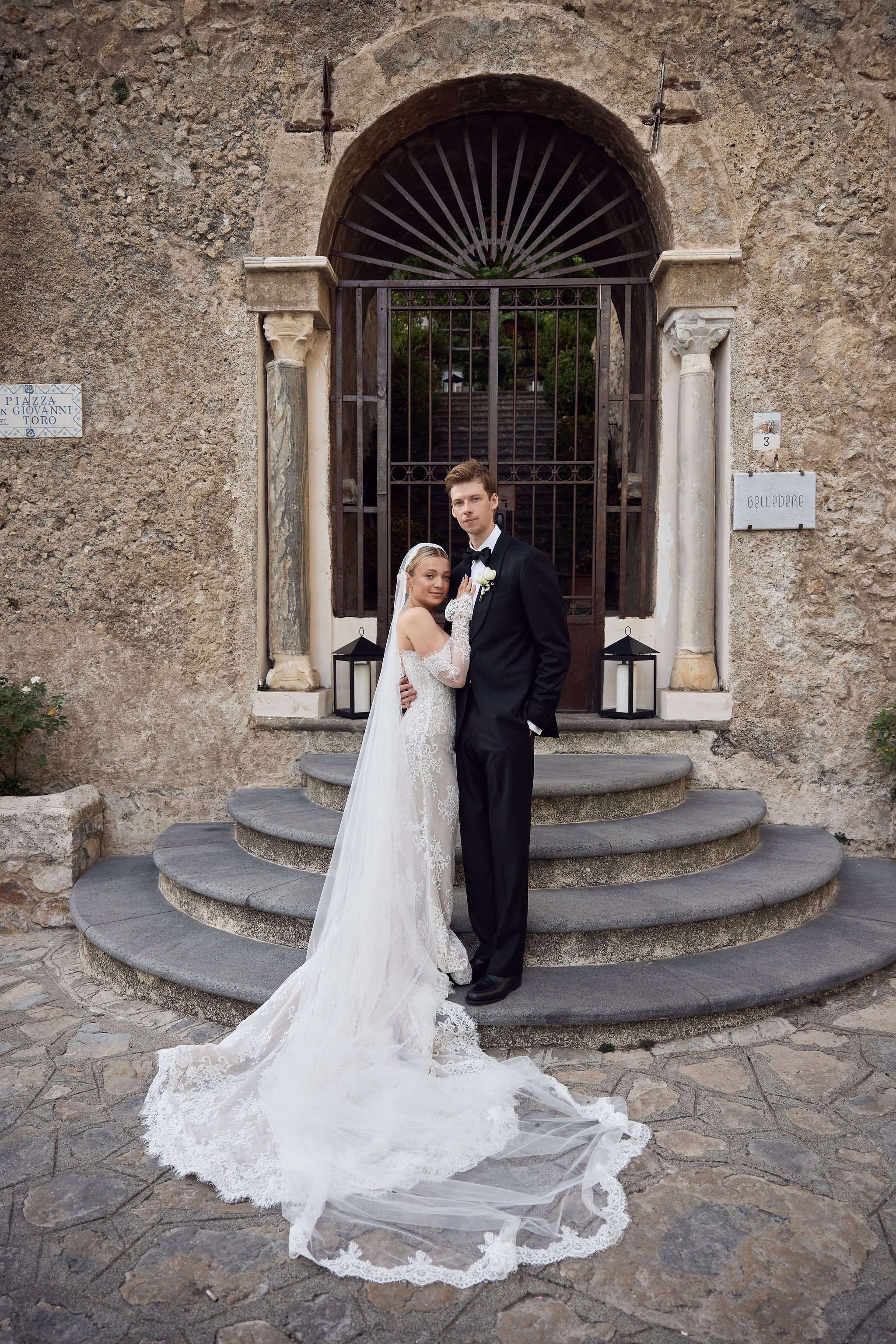 Bride and groom in wedding attire standing on stone steps in front of an old stone wall with a gated archway, holding each other romantically.