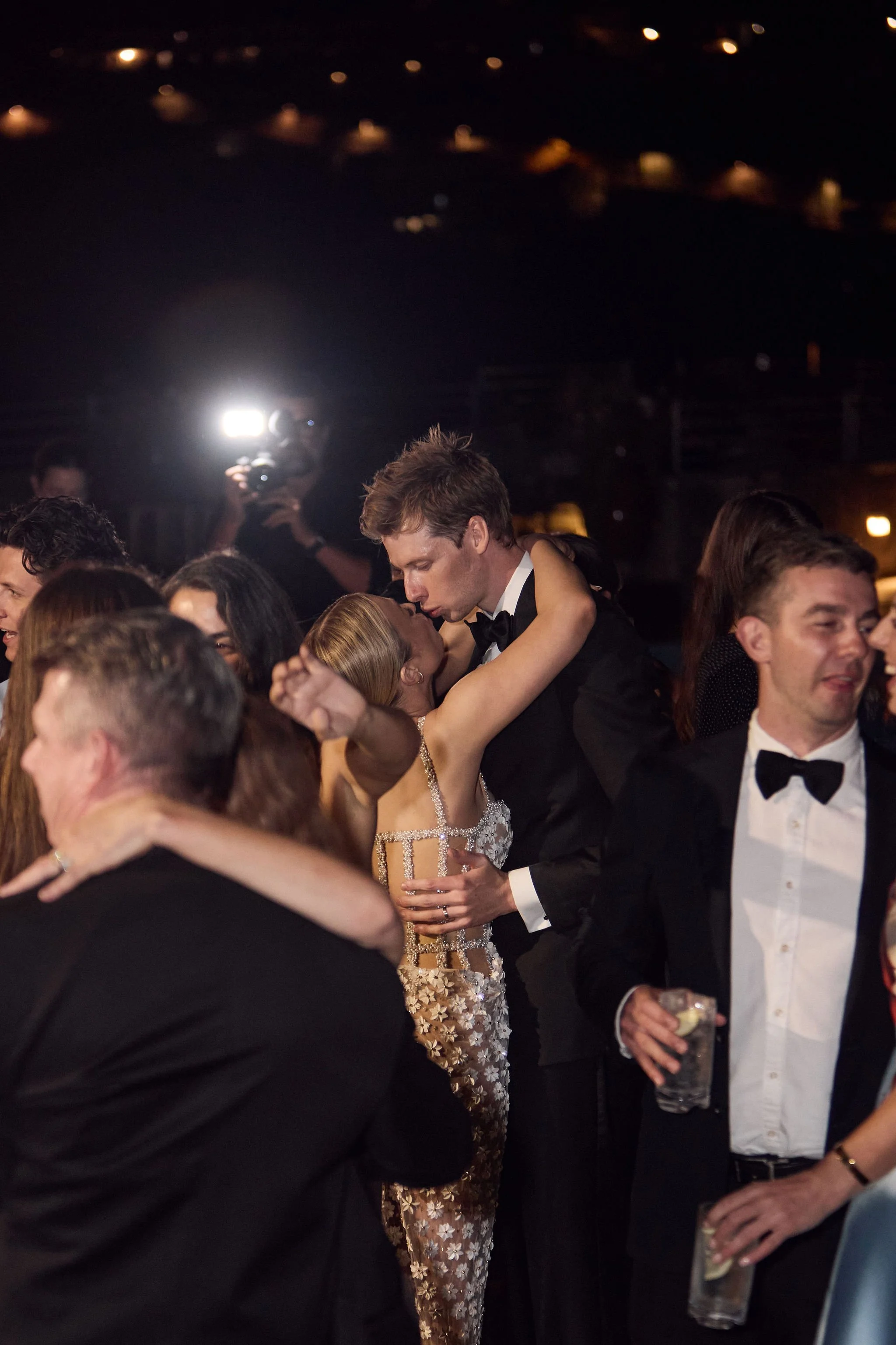 A couple in formal attire kissing at a dance floor while surrounded by other dancing guests, with bright lights and a photographer in the background.