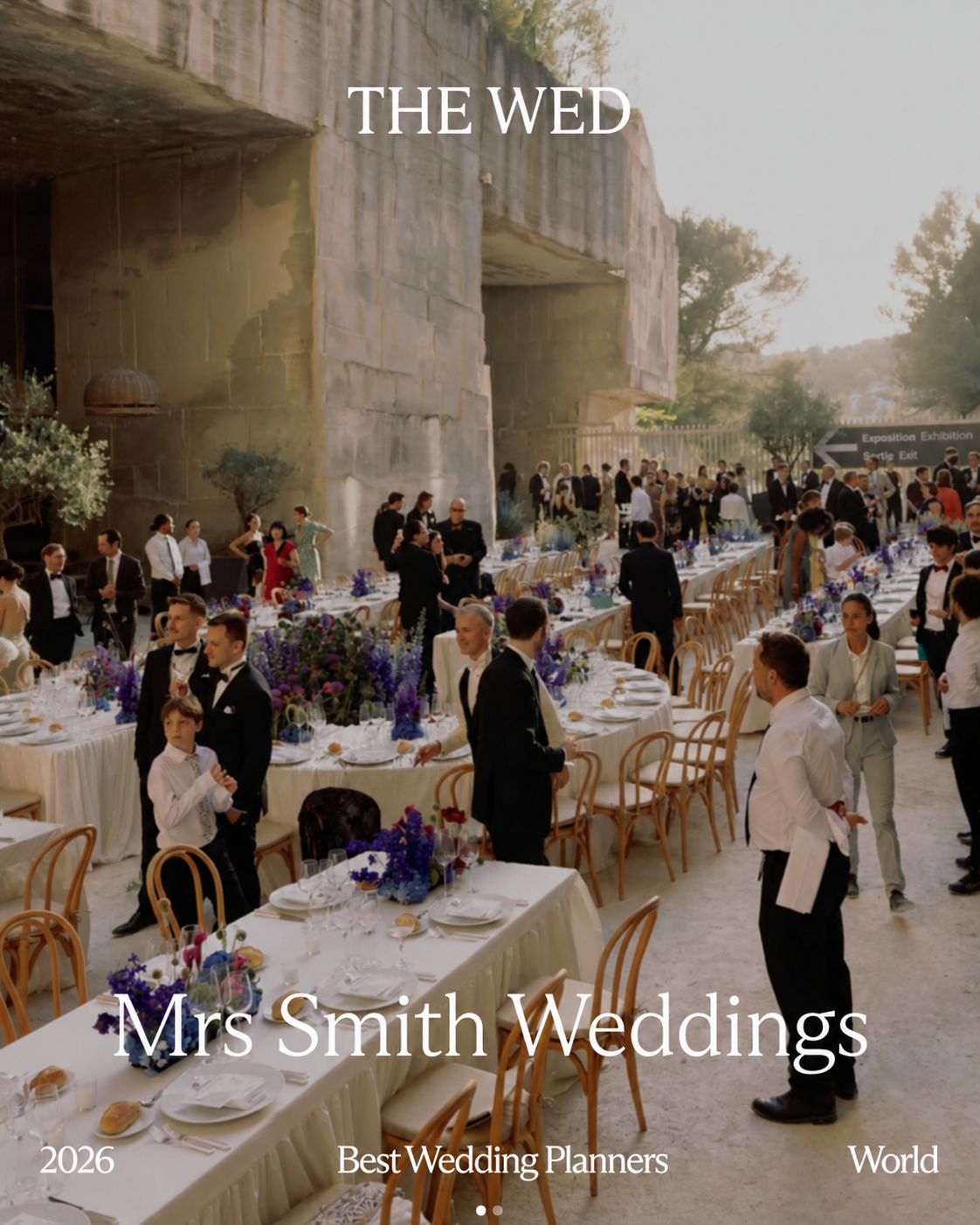 Guests at an outdoor wedding reception with purple floral arrangements and a building in the background.