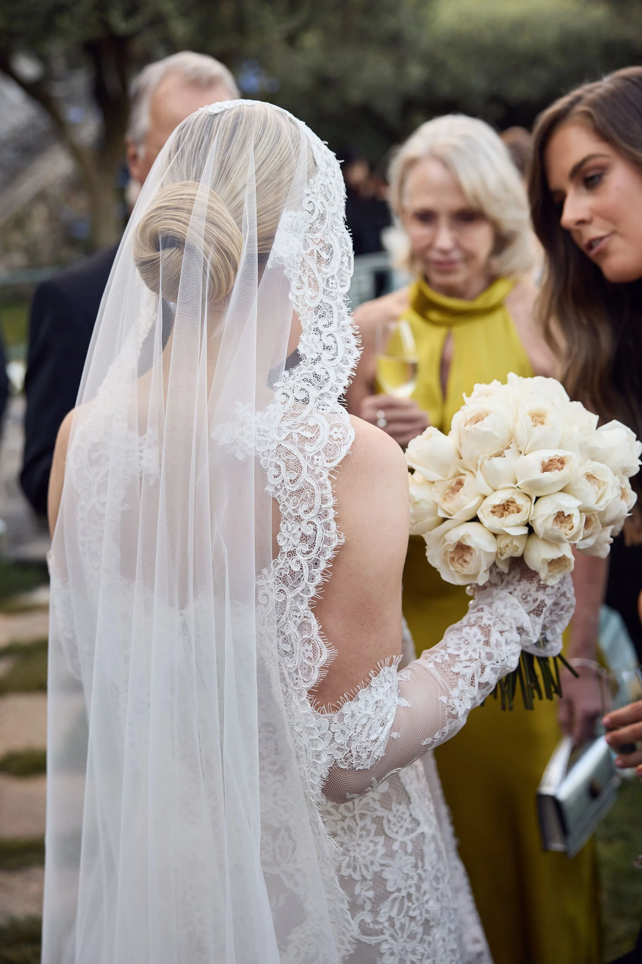 A bride in a lace wedding dress and veil holds a bouquet of white roses at an outdoor wedding reception.
