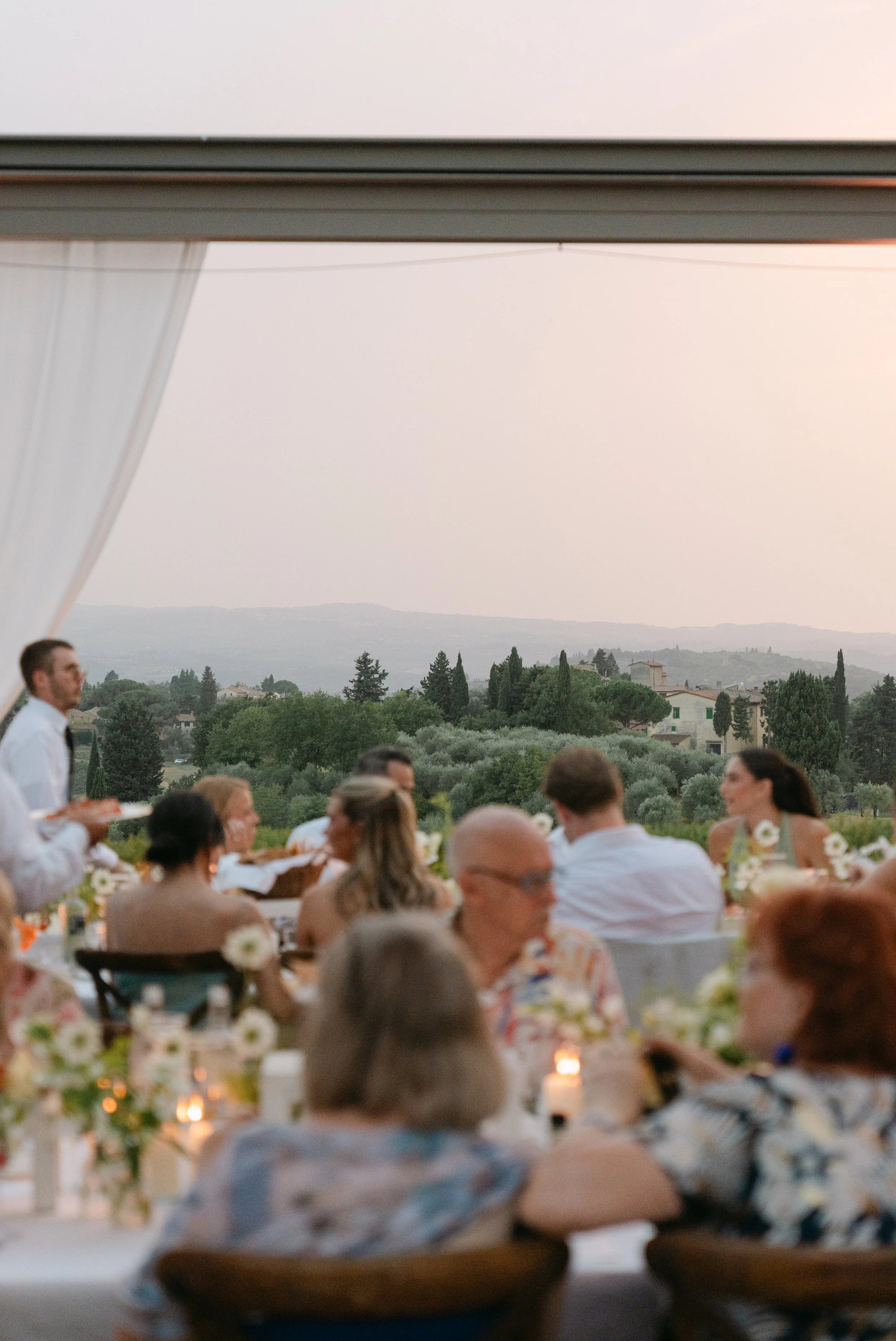 People dining outdoors during sunset with a scenic view of trees and hills in the background.