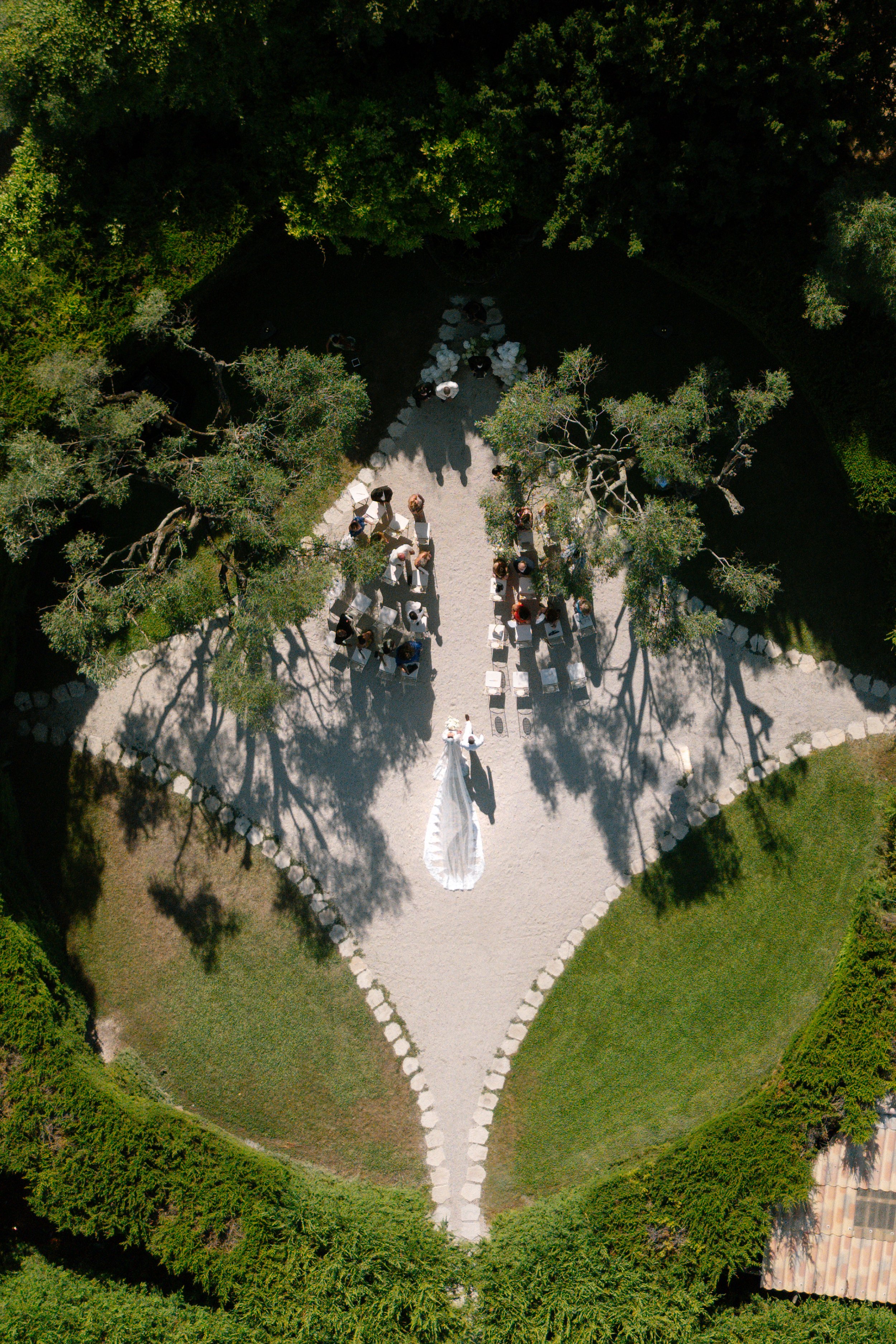 Aerial view of a wedding ceremony setup in a scenic outdoor garden with green trees surrounding a gravel aisle and white chairs.
