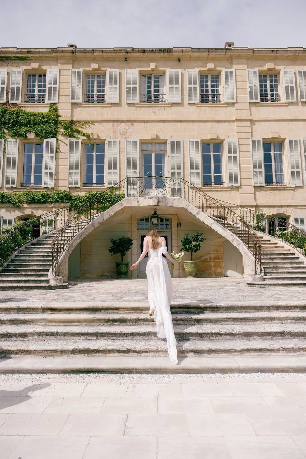 A woman in a white wedding dress holding a bouquet standing on stone steps in front of an elegant building with multiple windows and white shutters.