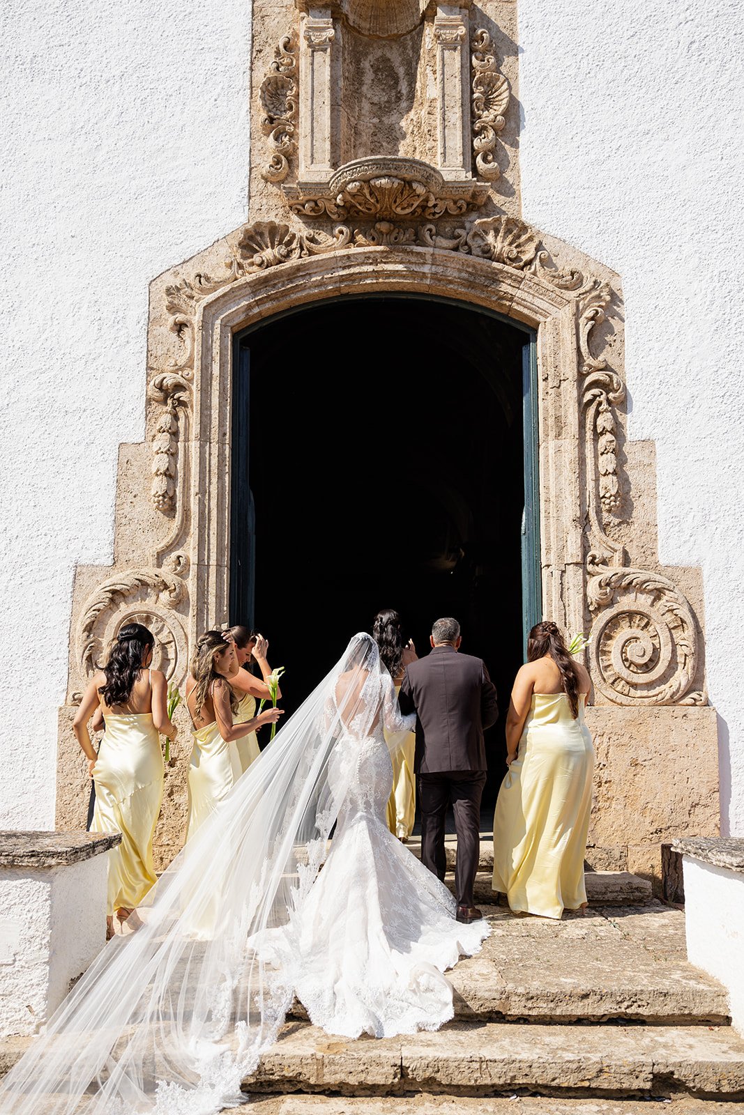 A bride in a white wedding gown with a long train and veil stands with her back to the camera, holding a bouquet, as she enters a building with a group of bridesmaids in yellow dresses and a man in a dark suit.