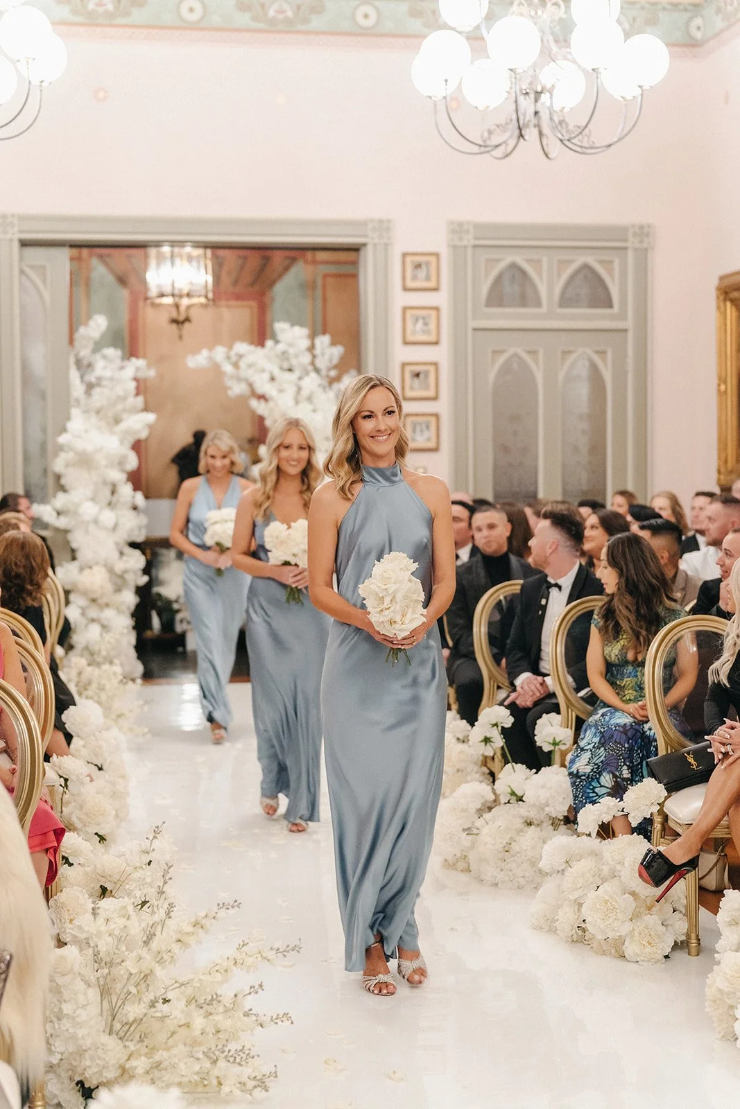 Bridesmaids walking down the aisle at a wedding ceremony, holding white bouquets, in a decorated venue with white floral arrangements and chandeliers.
