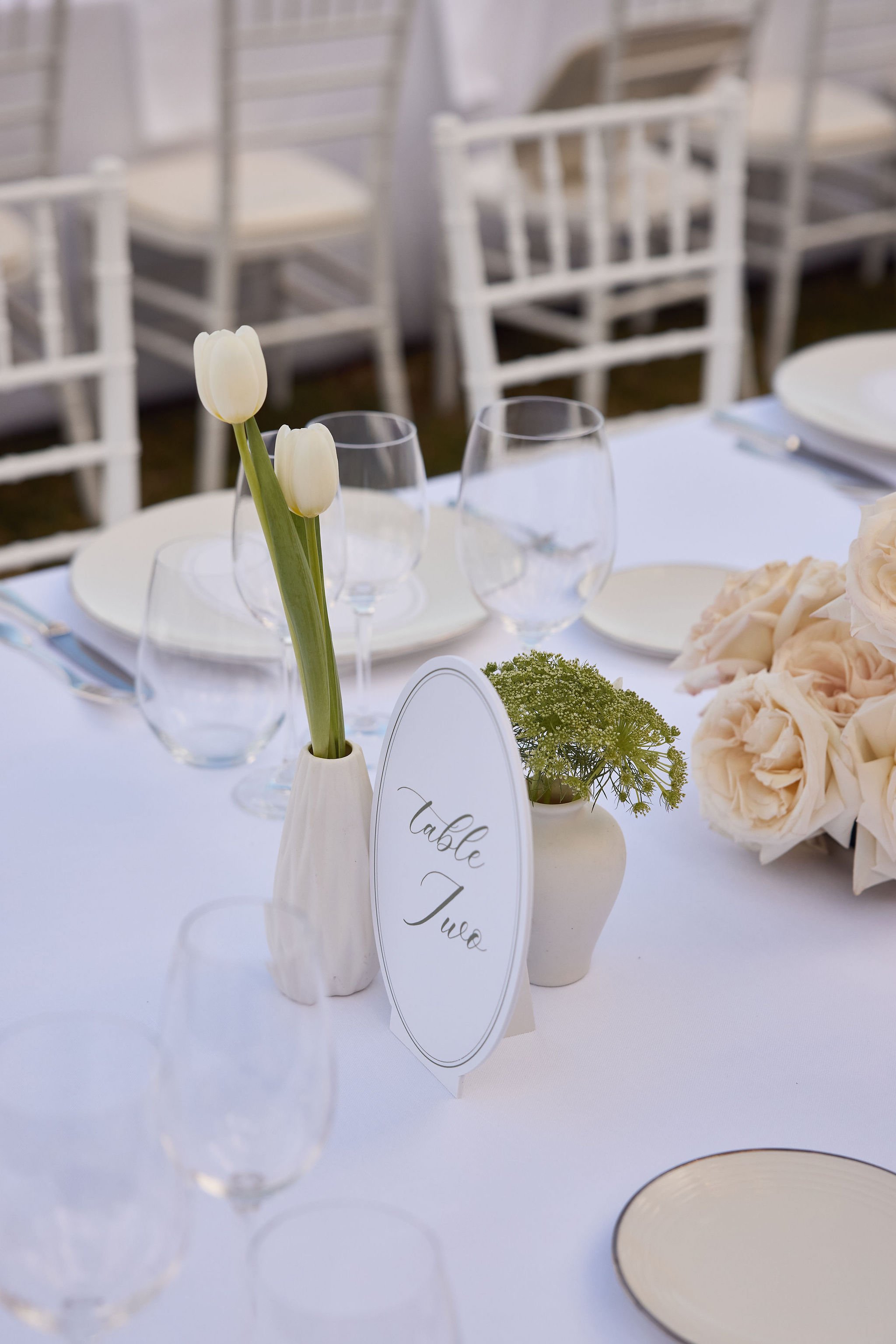 A table setting with white tablecloth, glassware, white plates, and floral centerpieces, including white tulips and roses, with a table sign labeled 'Table Two' in elegant script.