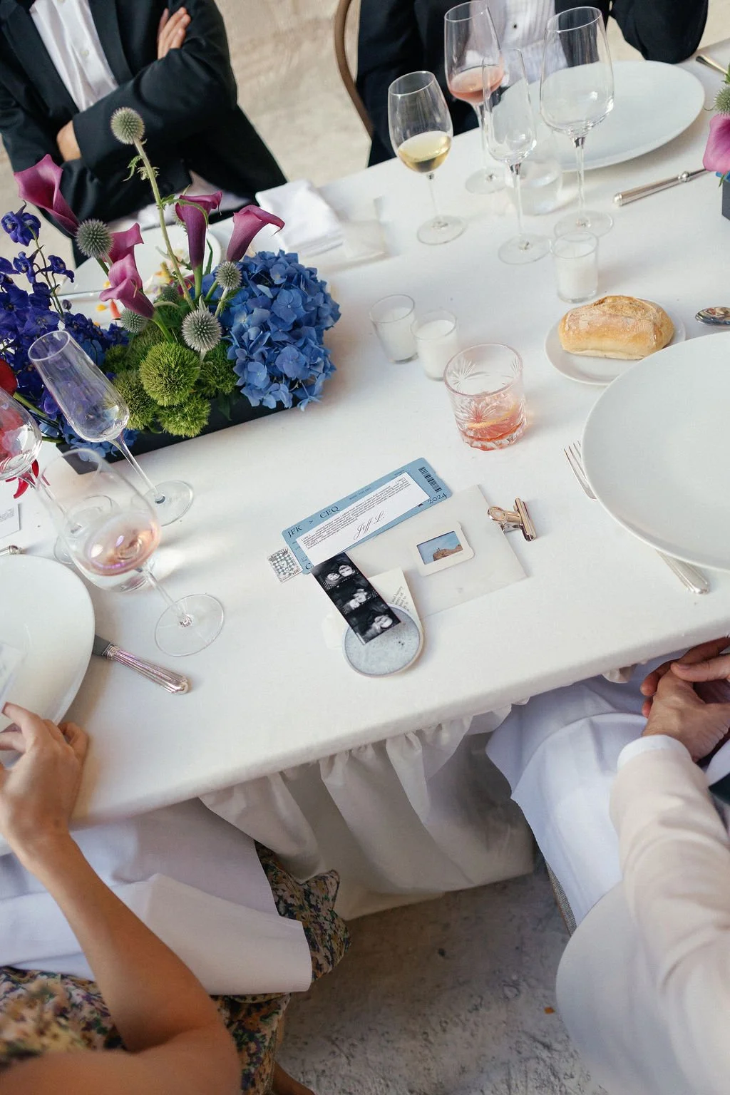 A formal dining table set with wine glasses, a floral centerpiece, a bread roll, a ticket, and a few small candles, surrounded by people in elegant attire.